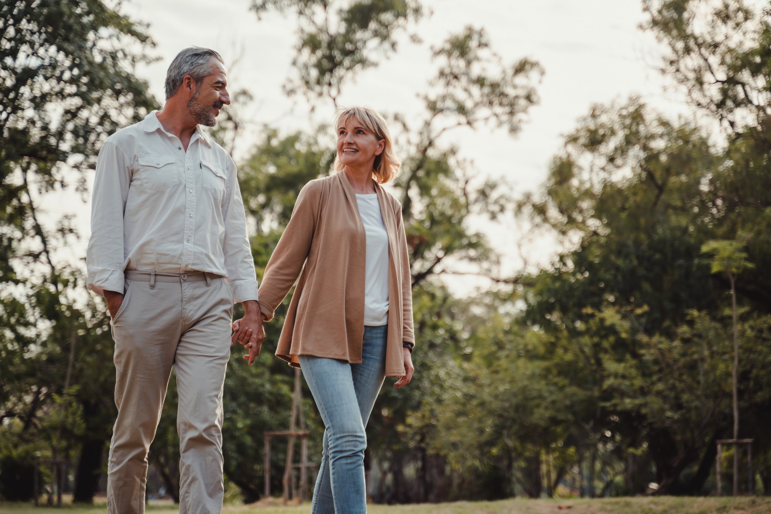 A middle-aged couple walking hand-in-hand in a park surrounded by trees