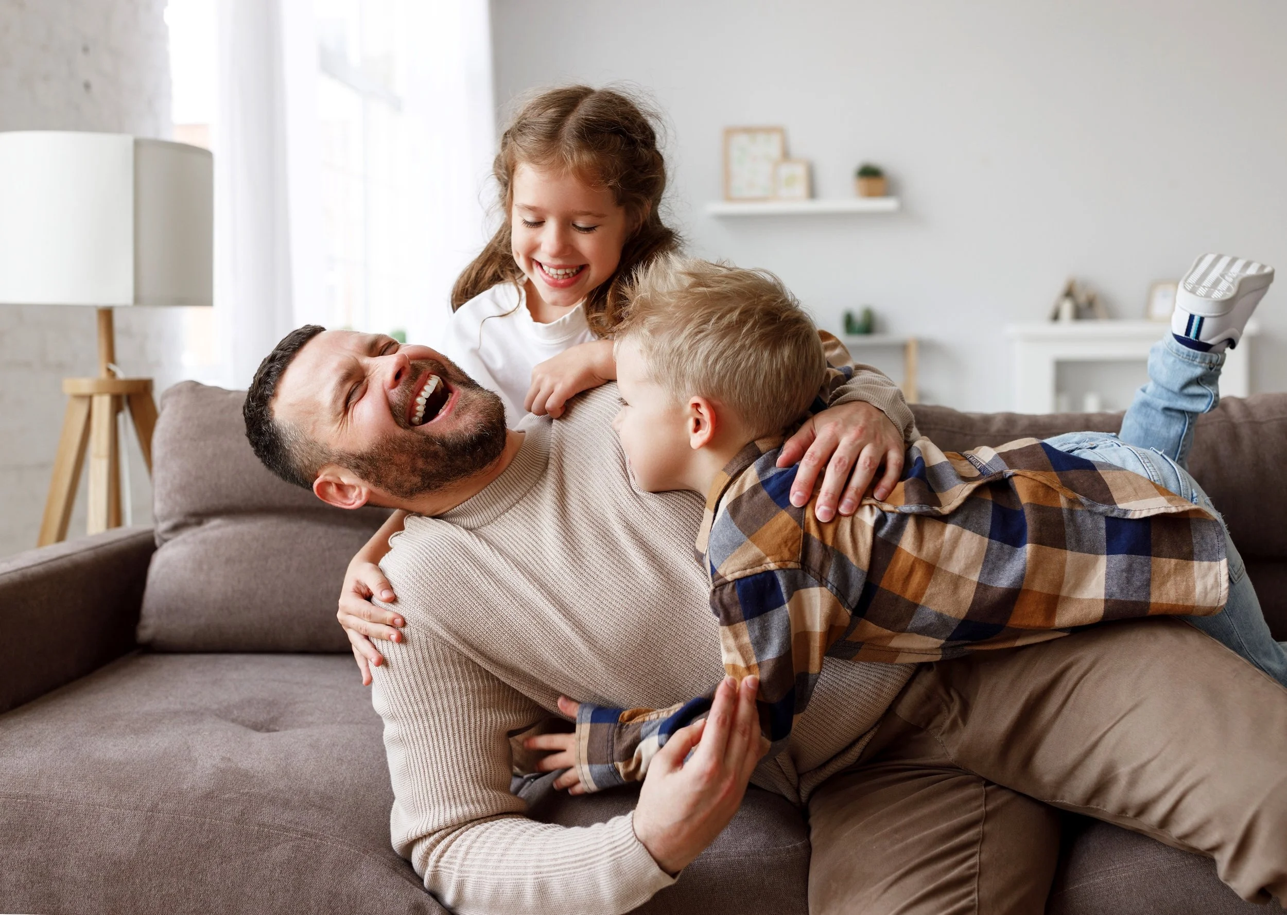 A man with a beard laughs as two children playfully climb on him on a sofa in a bright living room.