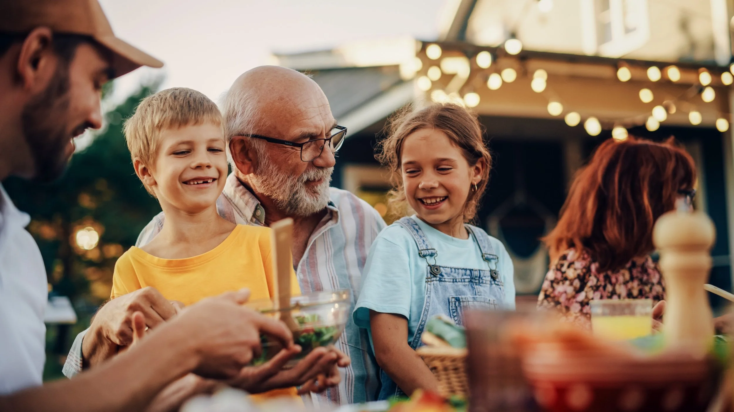 A family gathered outside at a table enjoying food and conversation, with string lights and trees in the background.