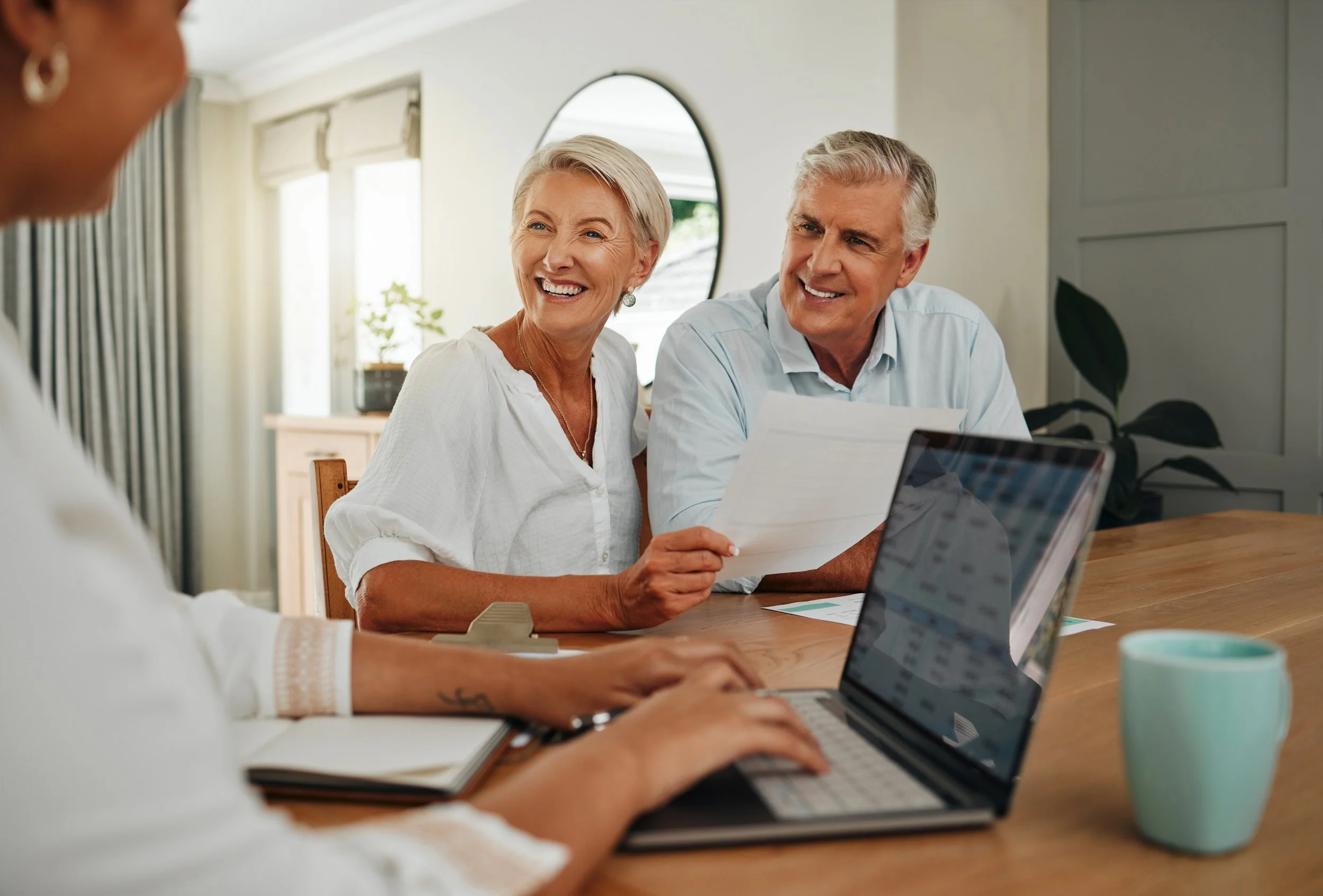 Smiling elderly couple discussing financial documents with advisor during meeting at home, with open laptop and coffee mug on table.