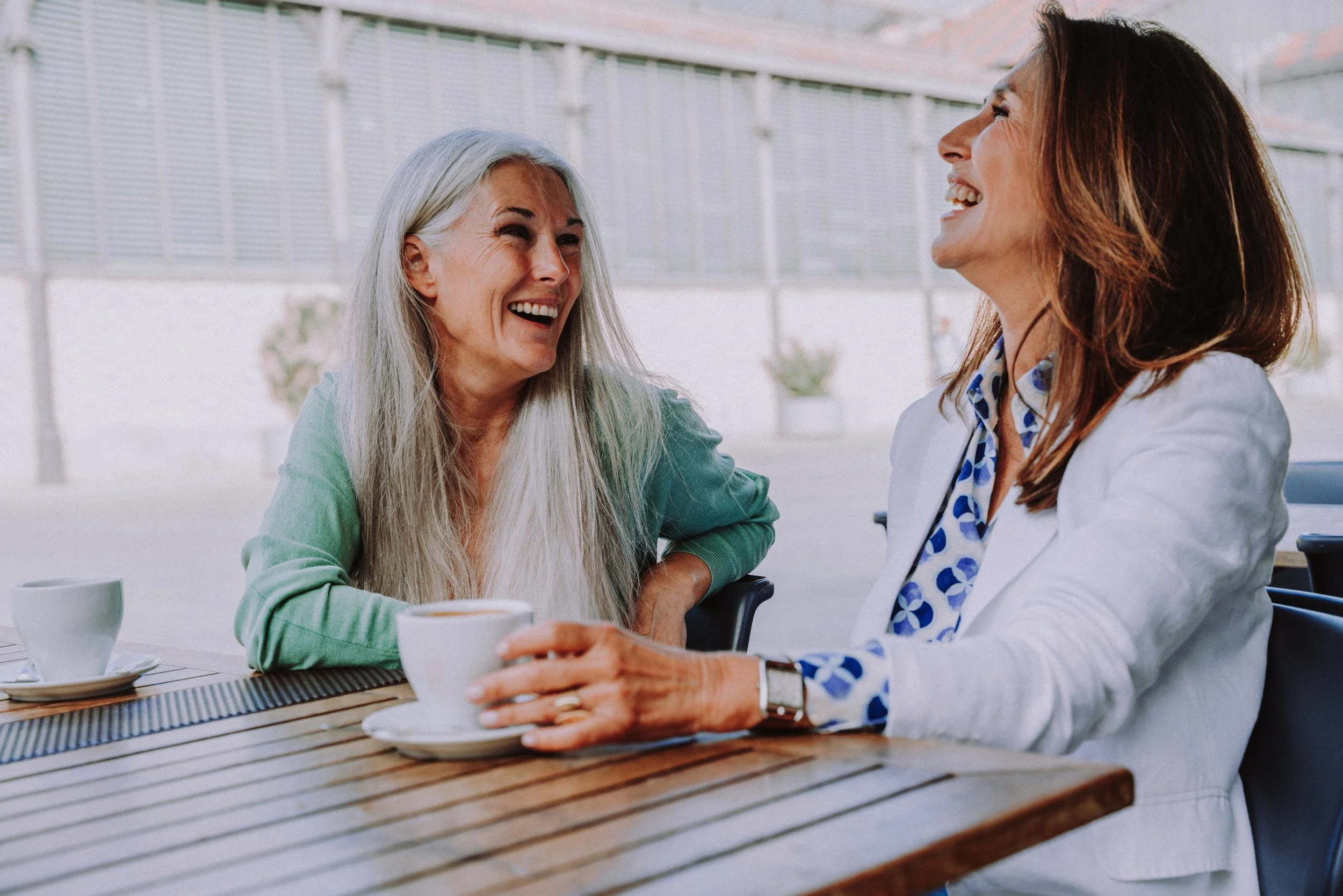 Two women sitting at a wooden table, sharing a laugh, in a bright, modern outdoor cafe, with cups of coffee in front of them.
