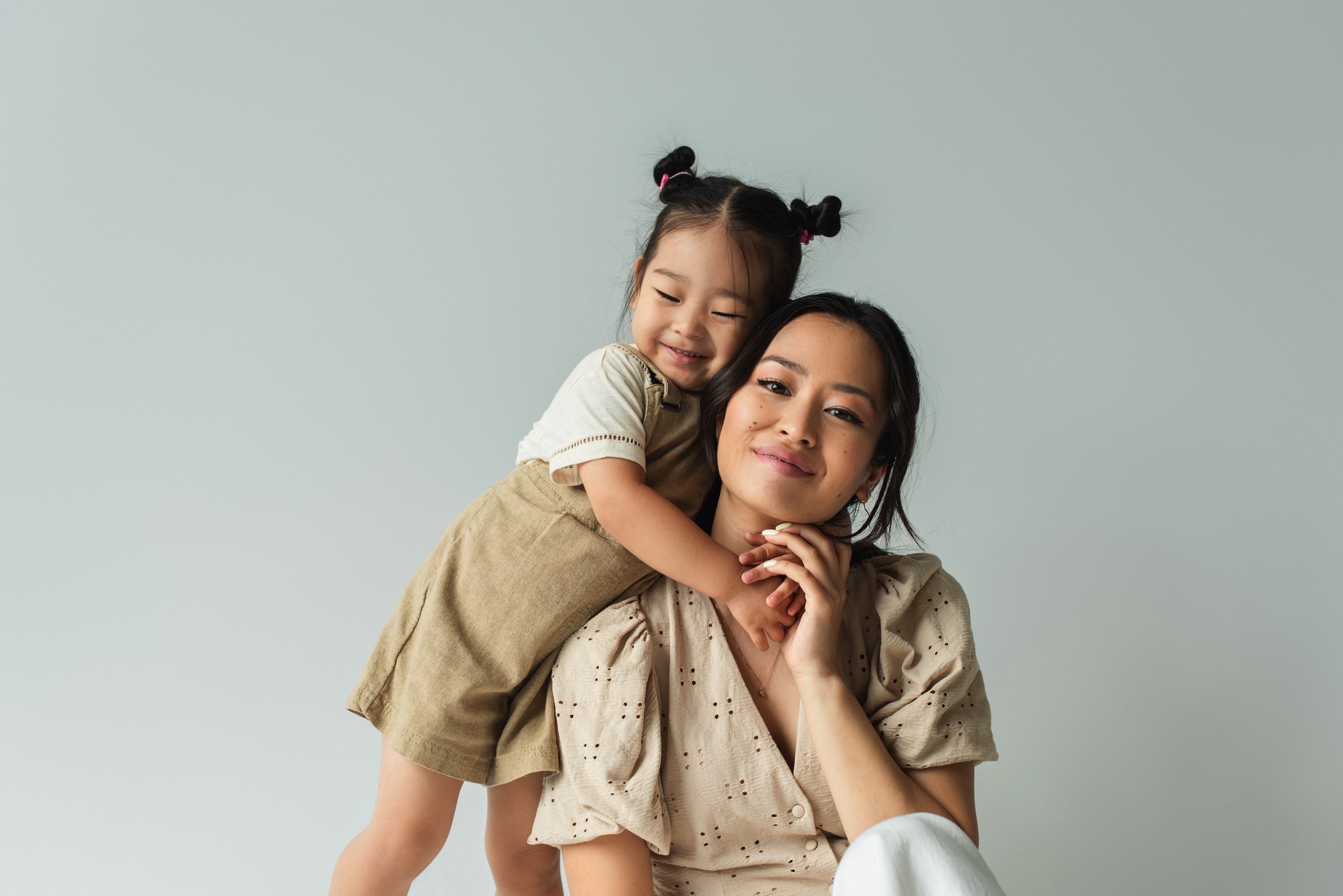 A woman with dark hair, wearing a beige blouse, holds her chin with her hand while a young girl with pigtails, wearing a beige dress, hugs her from behind with a smile. They are against a plain light background.