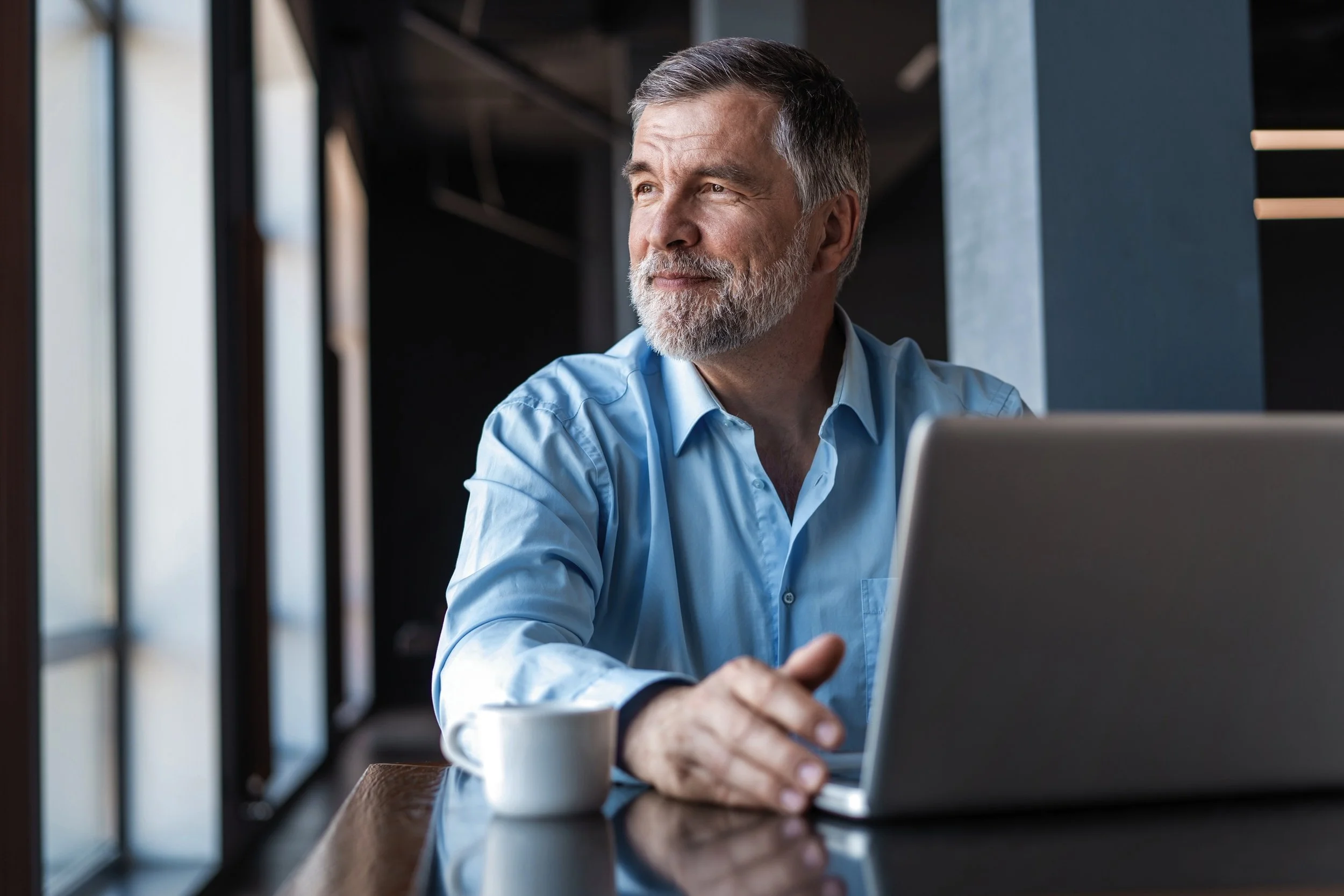 A middle-aged man with gray hair and beard, wearing a light blue shirt, sitting at a table with a laptop and a white coffee mug, looking thoughtful and gazing out a window in a modern office setting.