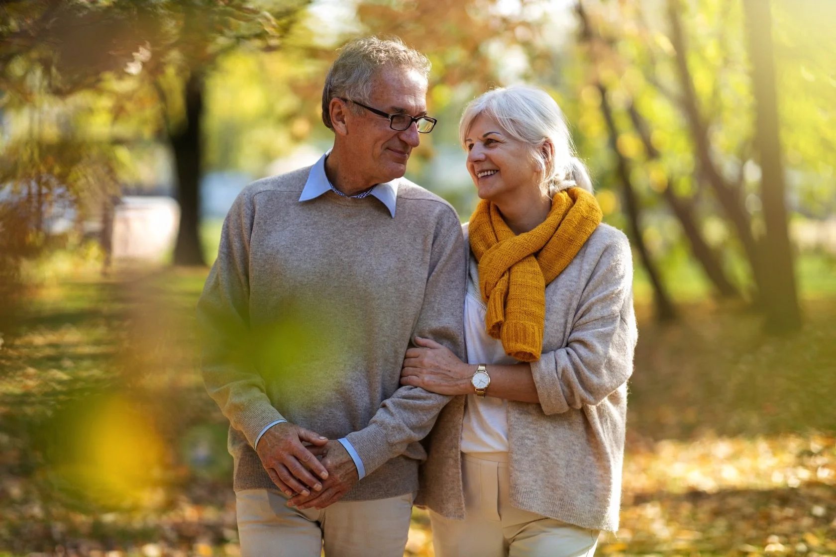 An elderly couple walking together outdoors in a park during autumn, sharing a tender moment as they look into each other's eyes, surrounded by fall foliage.