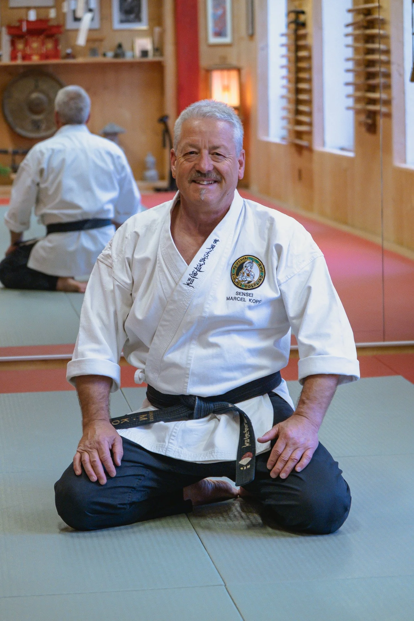Ein älterer Mann in einem Karateanzug sitzt in der Mitte auf den Knien auf Tatami-Matte in einem Dojo, lächelt in die Kamera, im Hintergrund ein weiterer Mann in Karate-Kleidung, japanische Wanddekoration und Holzelemente.