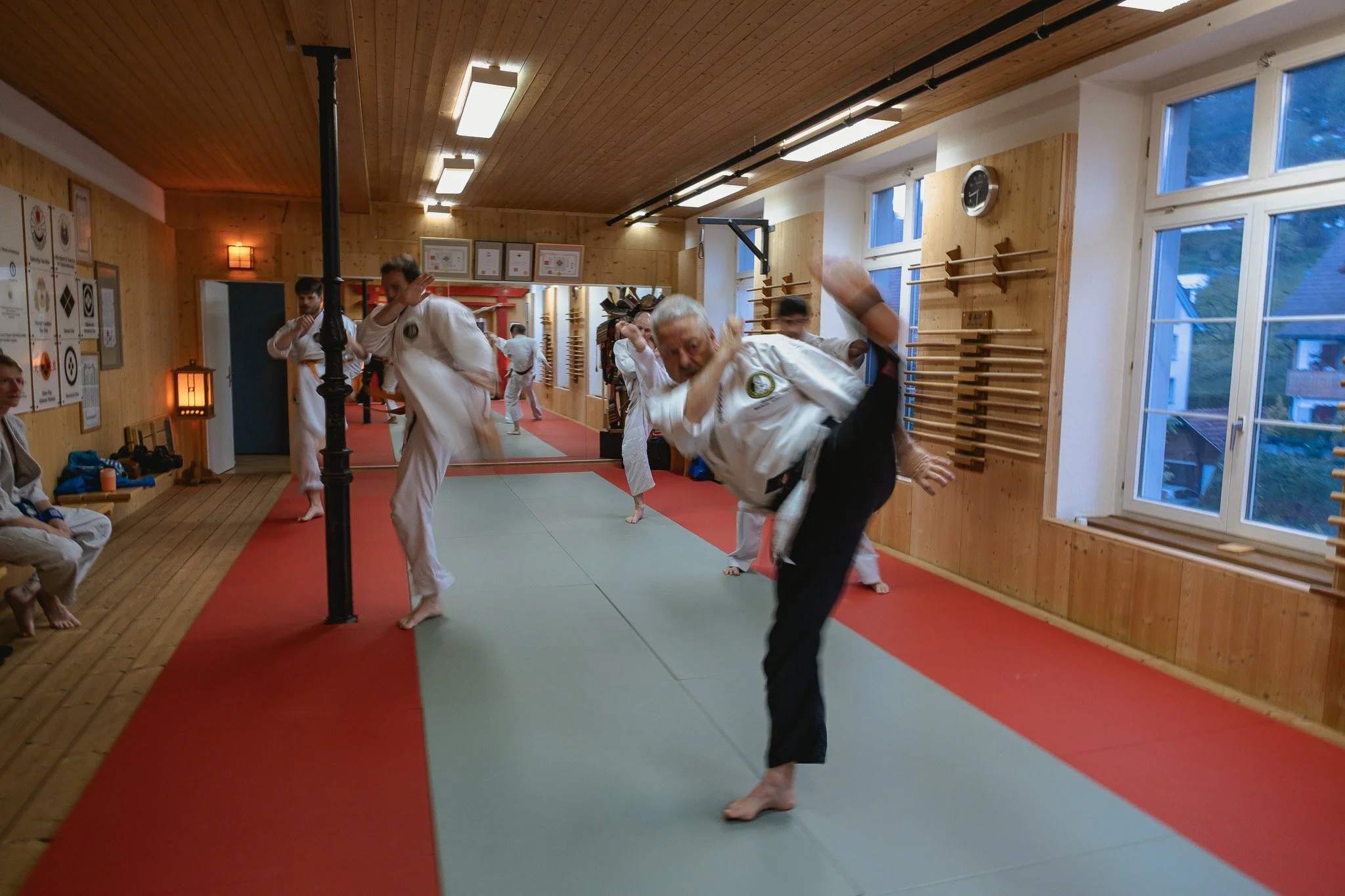 Menschen trainieren Taekwondo in einem Dojo aus Holz mit roten und grauen Matten auf dem Boden.