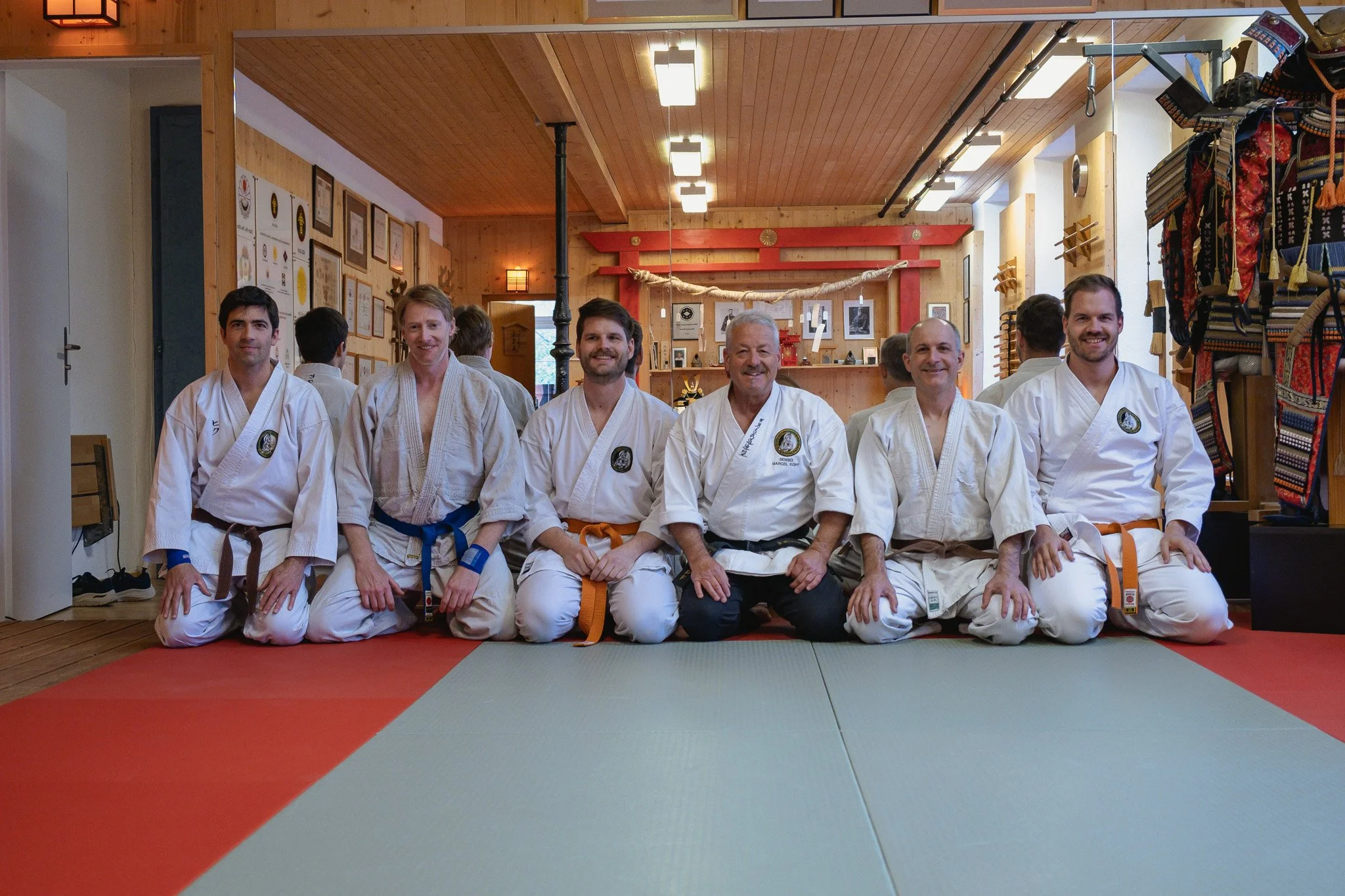 Gruppe von Selbstverteidigungs-Schülern in weissen Dōgi, sitzend mit ihrem Sensai auf einem Tatami-Matte in einem Dojo, mit Holz-Wänden und verschiedenen Bildern und Dekorationen im Hintergrund.