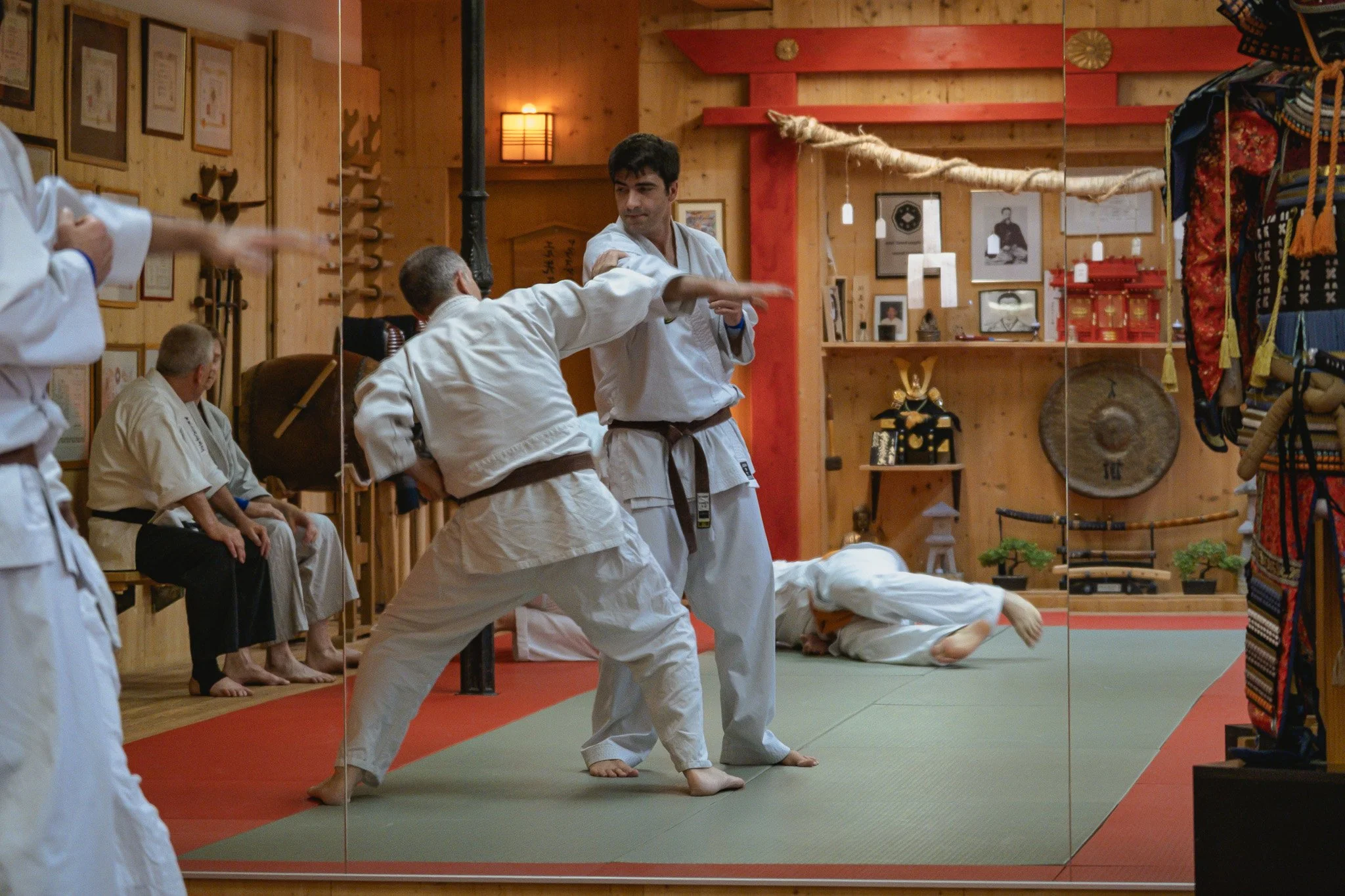 Zwei Männer üben gemeinsam in einem Dojo, der Raum ist mit Holz verkleidet und hat zusätzliche Dekorationen wie Bilder und Ausrüstungsgegenstände.
