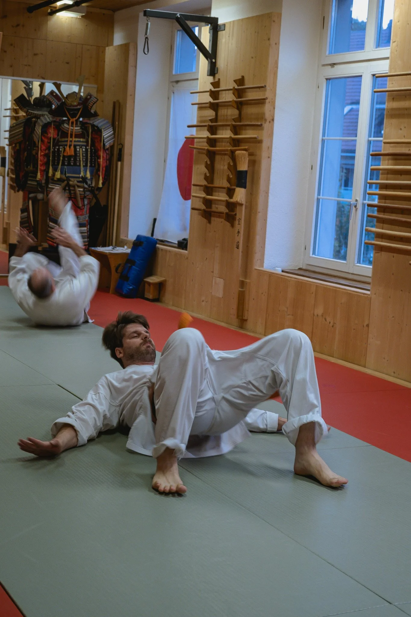 Zwei Männer trainieren Fallschule auf einer Tatami-Matte im Dojo, einer trägt ein weisser Dōgi, mit Holzregalen und einem Fenster im Hintergrund.