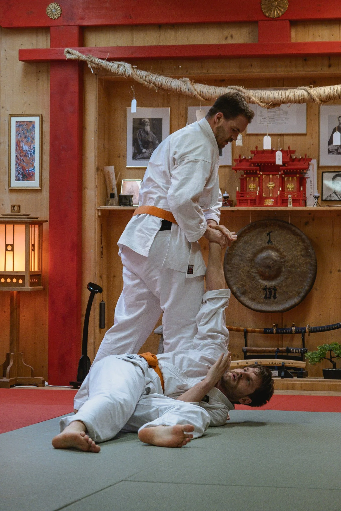 Zwei Personen in einem Kampfsporttraining, eine Person liegt auf dem Boden und die andere steht darüber, beide tragen weisse Dōgi. Im Hintergrund sind japanische Dekorationen und Bilder.