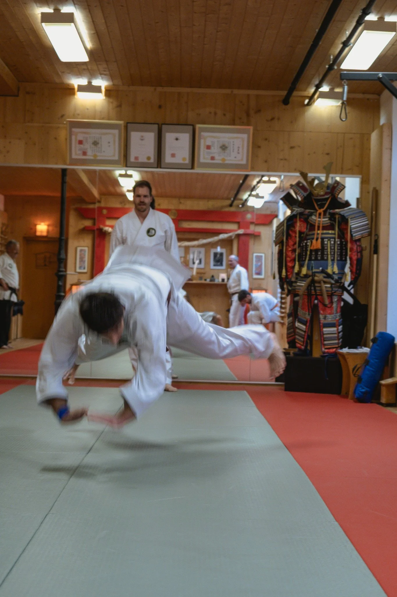 Junge im weissen Dōgi führt die sichere Fallschule aus, im Dojo mit Holzdecken und traditionellen Dekorationen.