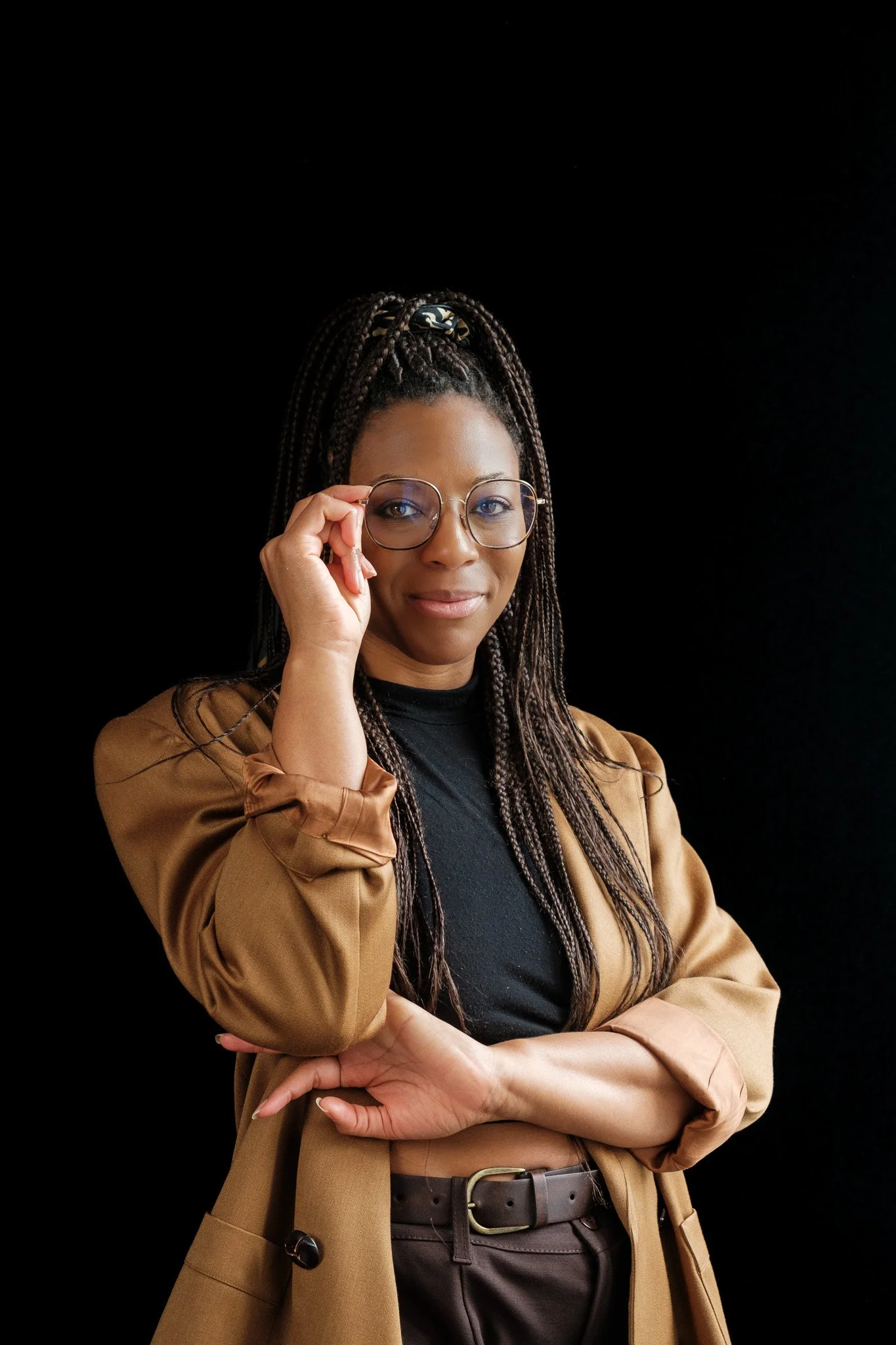 A woman with long braided hair wearing glasses, a black shirt, and a beige blazer, standing against a black background.