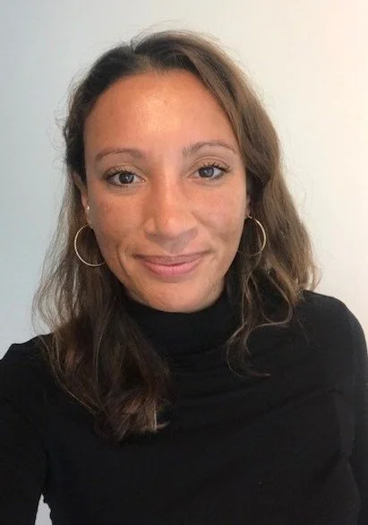 A woman with shoulder-length brown hair wearing a black turtleneck and hoop earrings, smiling at the camera against a plain white background.