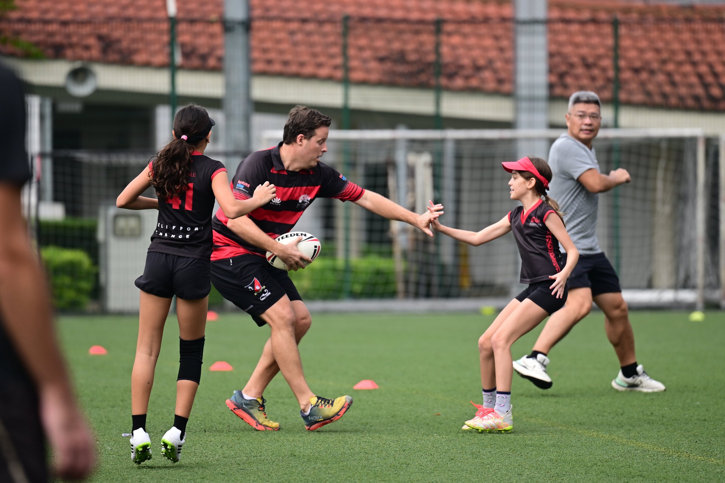 Two young girls playing rugby on a field, with an adult man in the background. The girls are wearing sports gear and are engaging in a game with the man, who is holding a rugby ball.