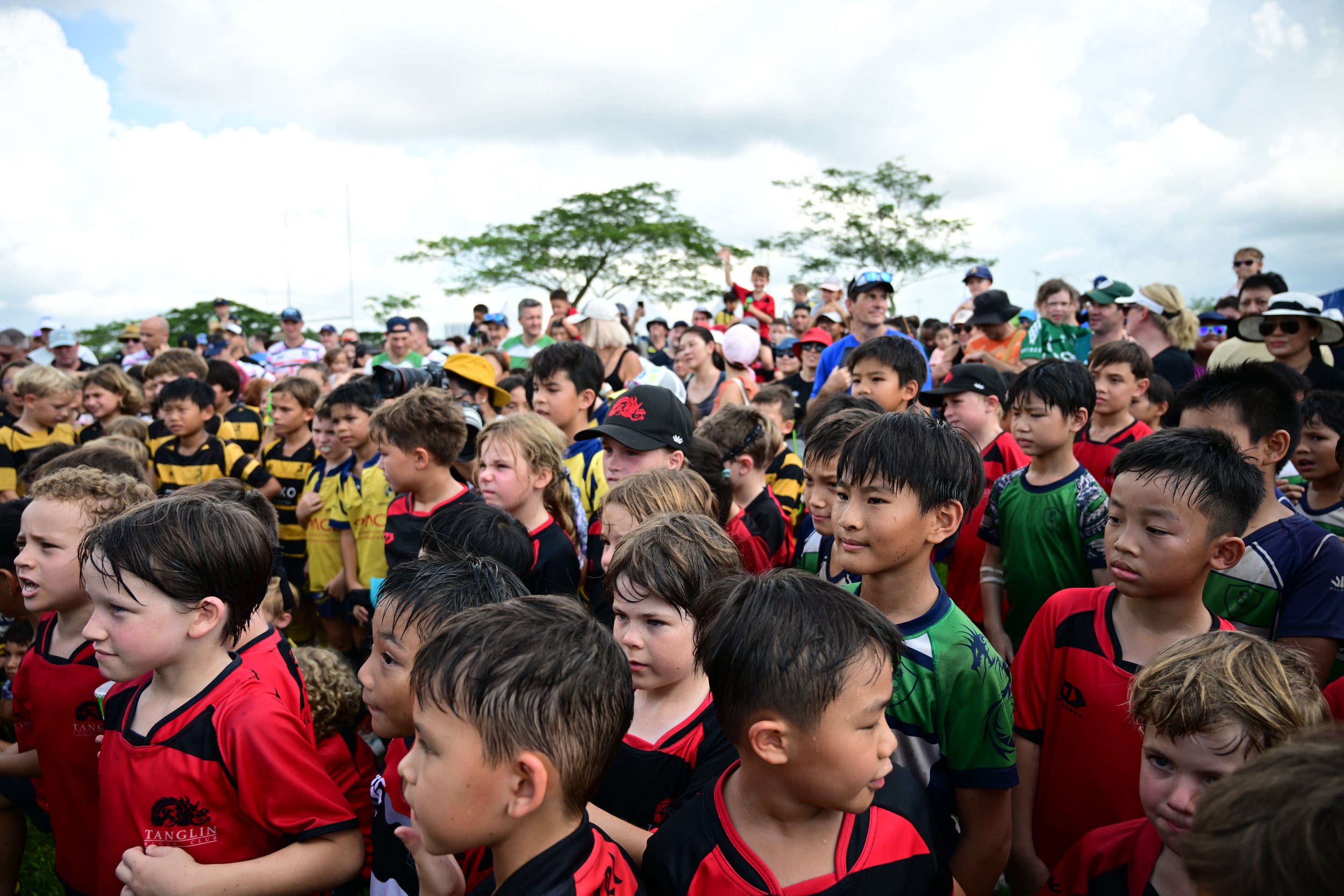 Group of children in colorful sports uniforms gathered outdoors, with adults and trees in the background.