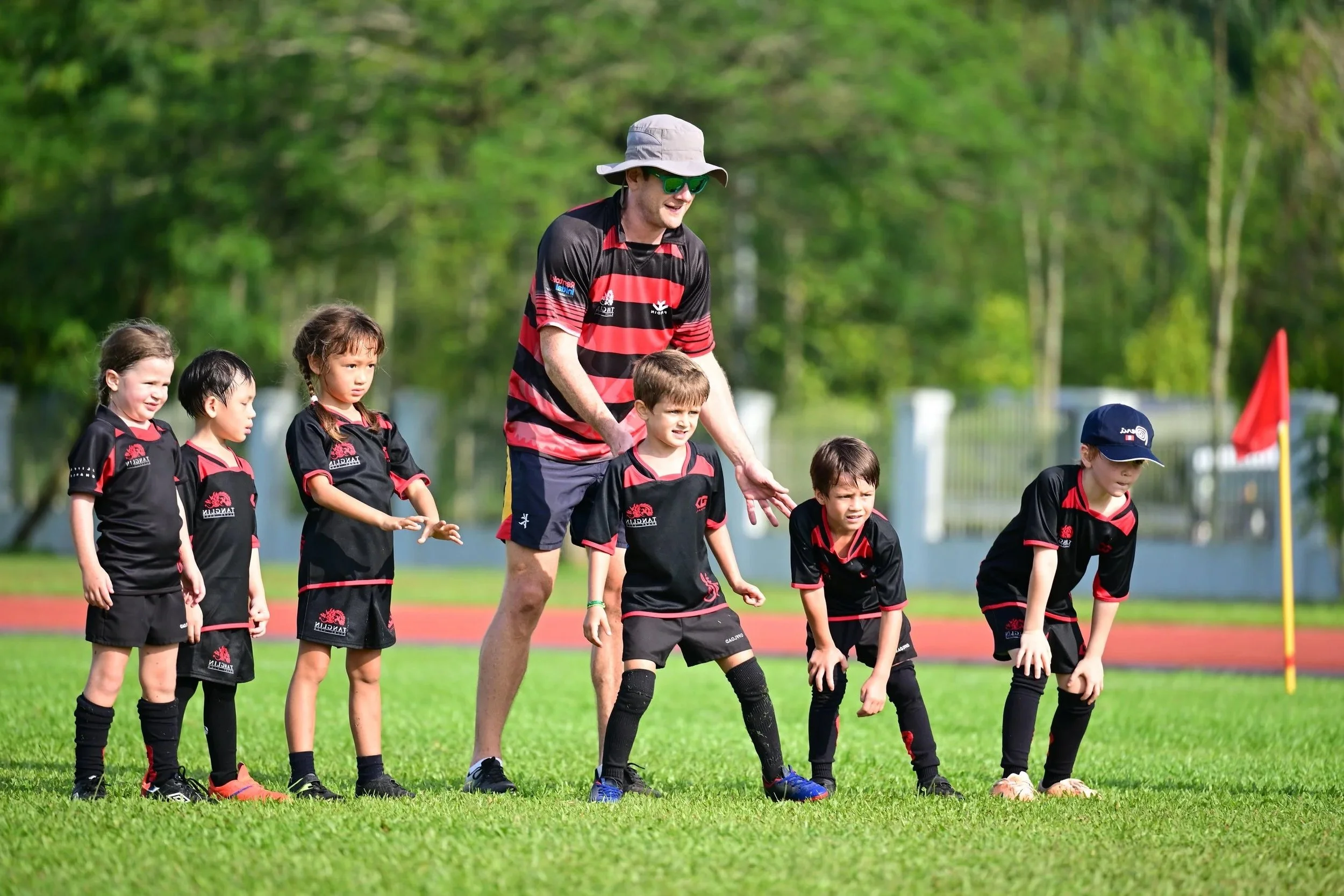 Children in black and red sports uniforms practicing rugby on a grassy field with their coach in a black and red jersey, green trees in the background, and a red flag on the side.
