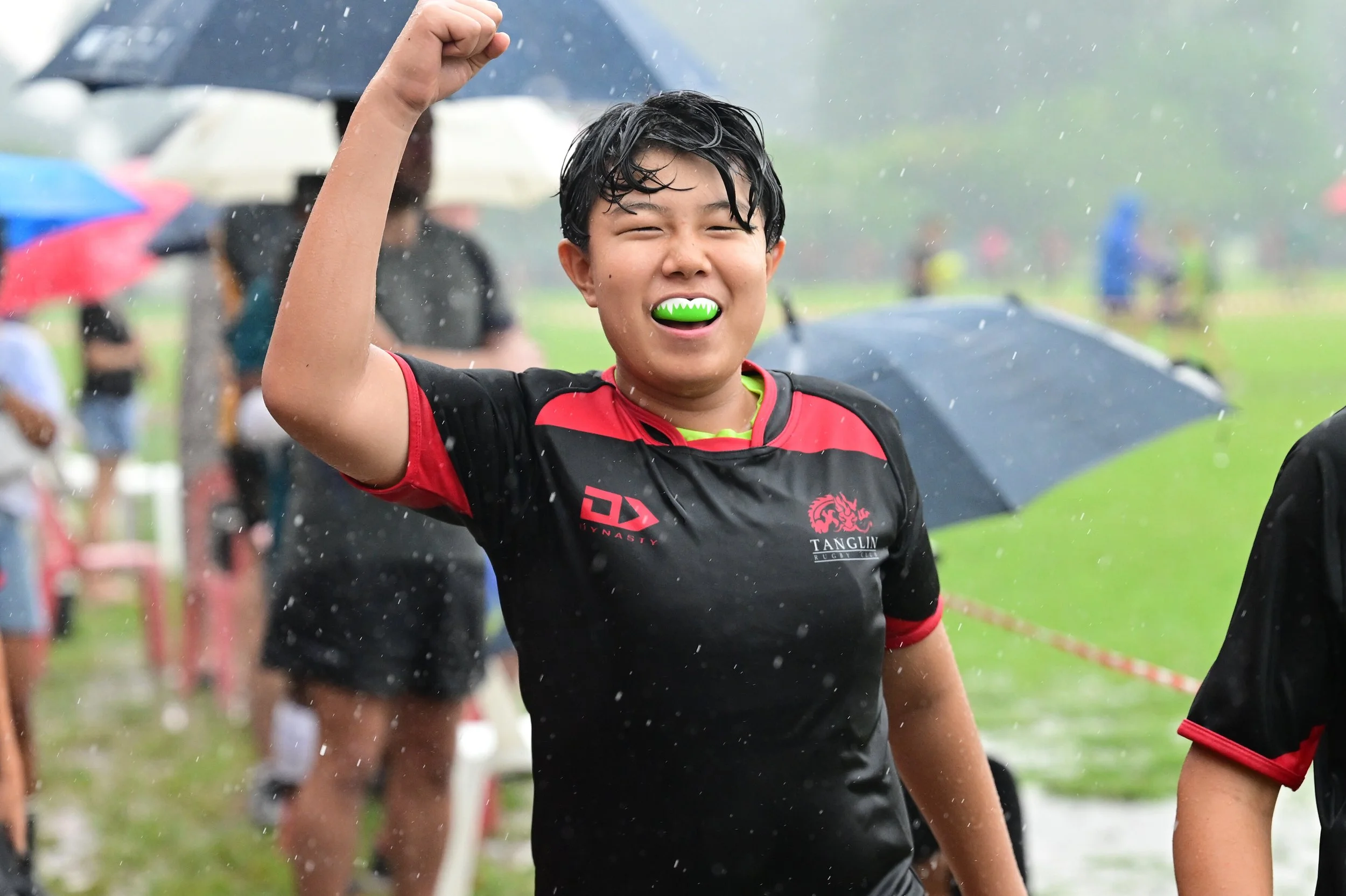 A young man smiling and raising his right fist in celebration during a rainy outdoor event, wearing a black and red sports jersey, with other people holding umbrellas and wearing rain gear in the background.