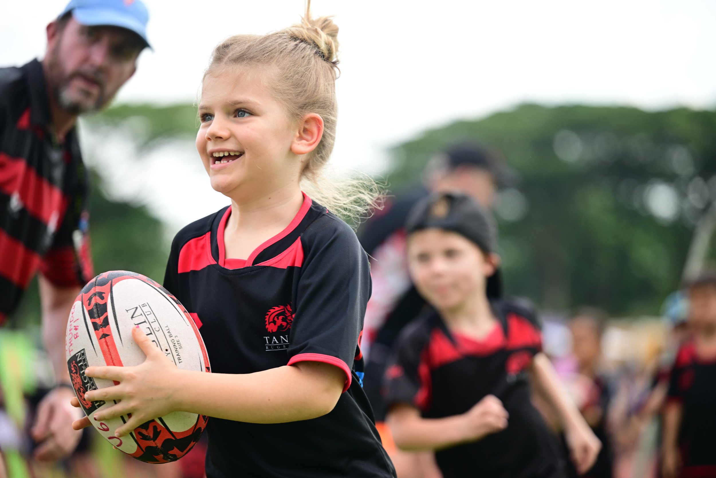 Young girl holding a rugby ball smiling during a youth rugby game, with other children and adults in the background.
