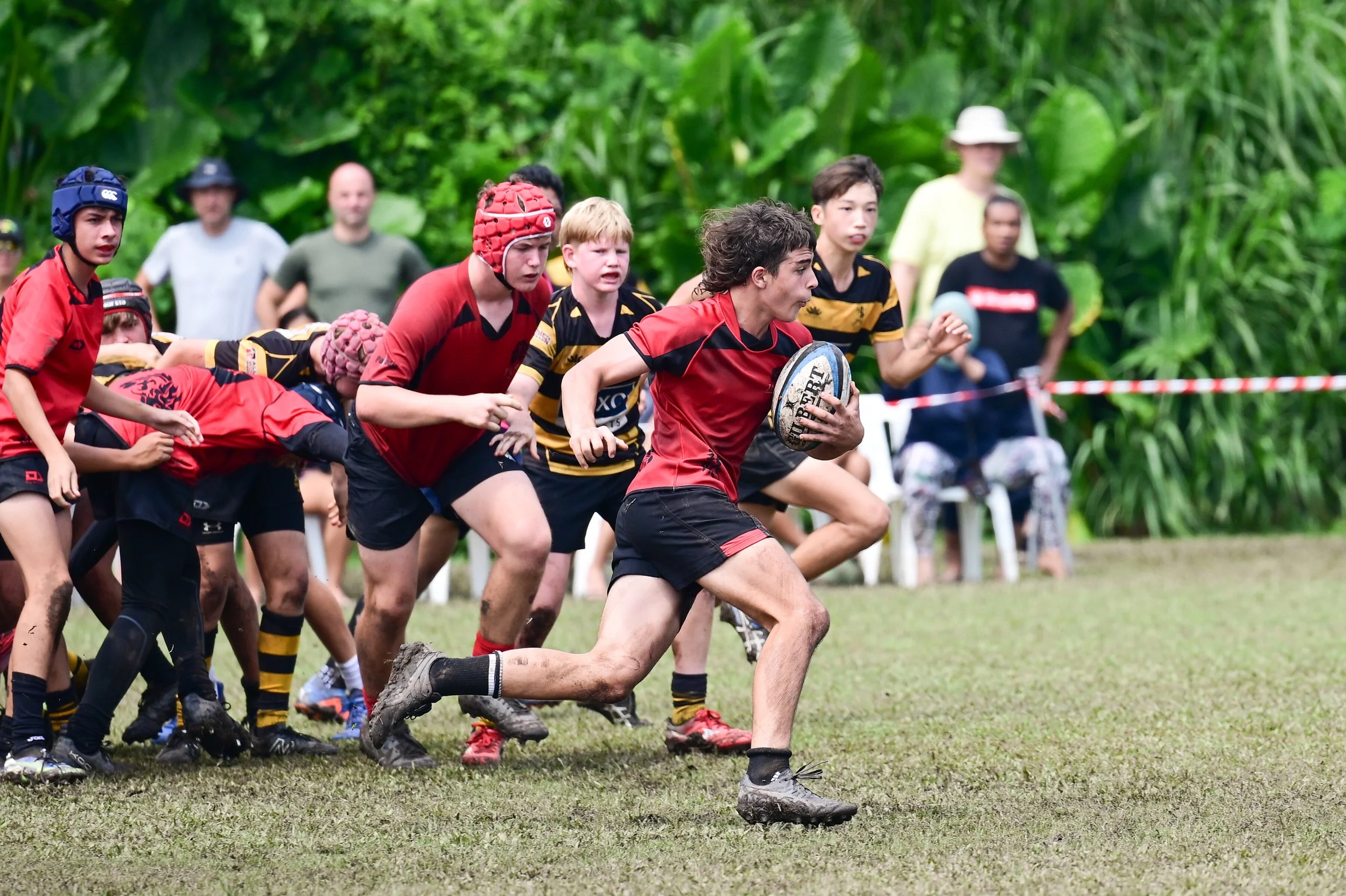 Rugby players in action on muddy field surrounded by lush green foliage; one player in red holding a rugby ball running forward, others attempting to block or tackle, with spectators in background