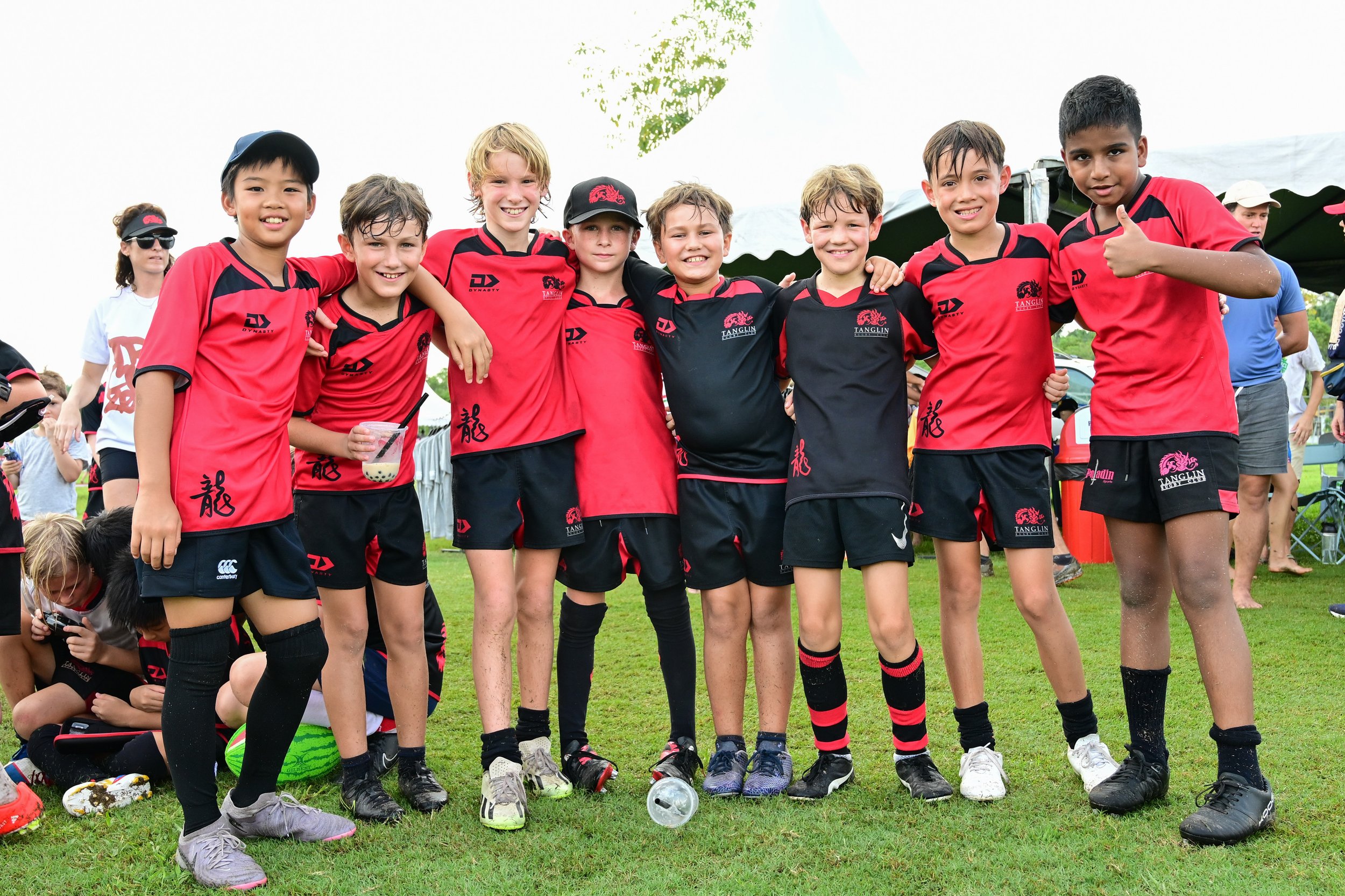 A group of young boys in red and black rugby uniforms standing arm in arm on a grassy field, smiling at the camera. They appear to have just finished a game or practice, with some holding drinks and others with wet hair.