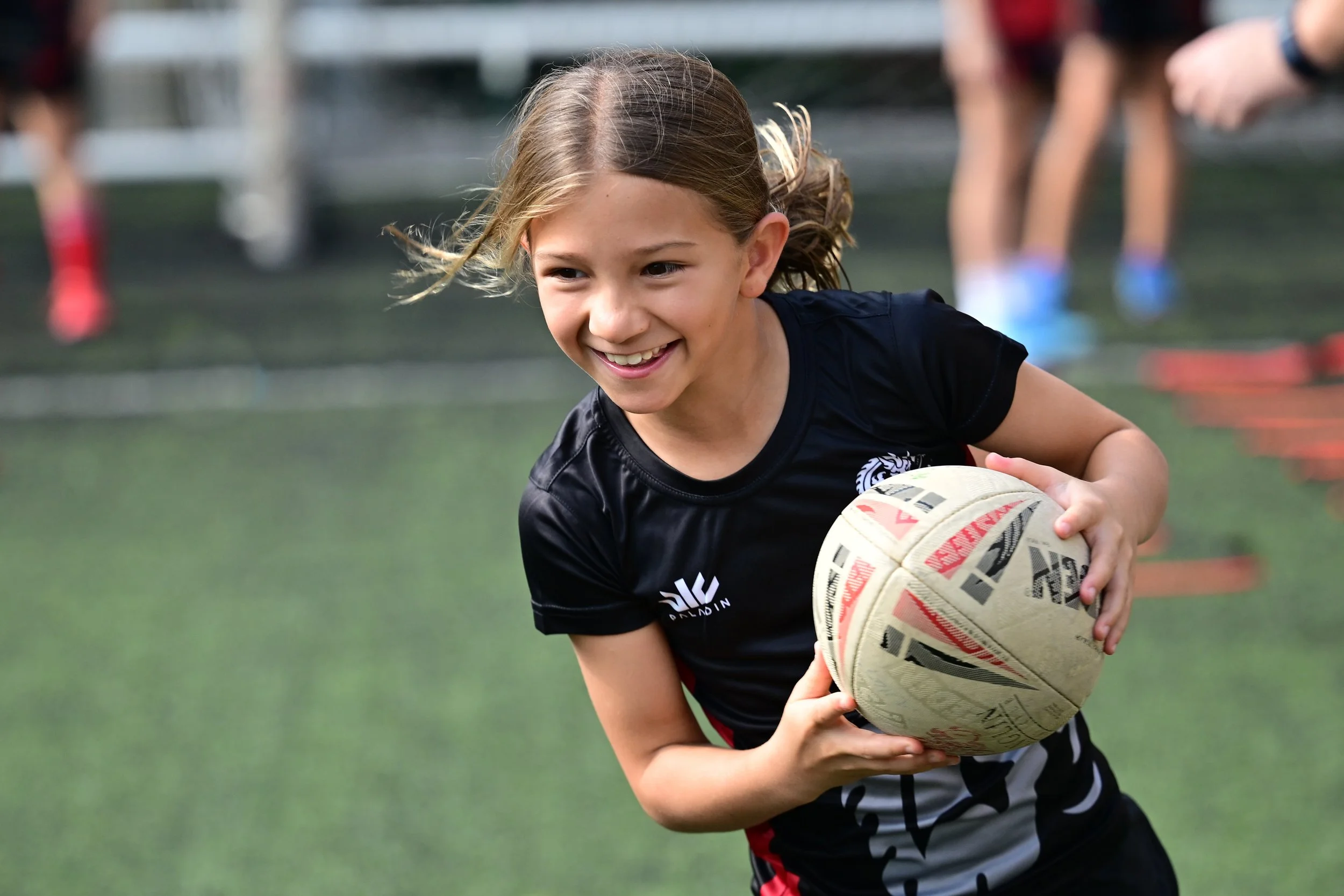 Young girl playing rugby on a field, smiling, holding a rugby ball, with other children in the background.