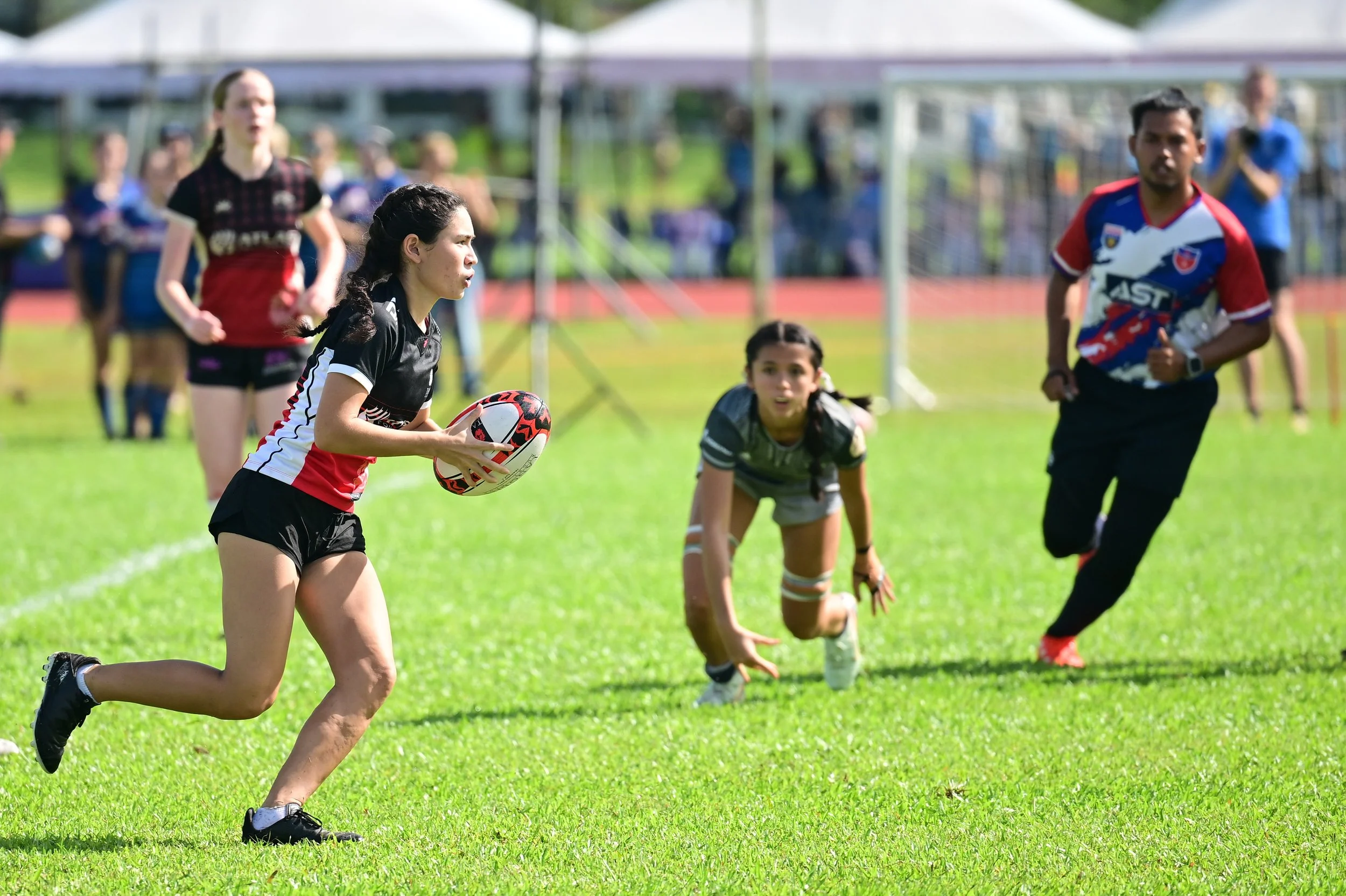 Women playing rugby on a grassy field during a match, with a woman in black and red holding the rugby ball, a woman in gray crouching, and a man in a colorful jersey running nearby, with spectators in the background under a tent.