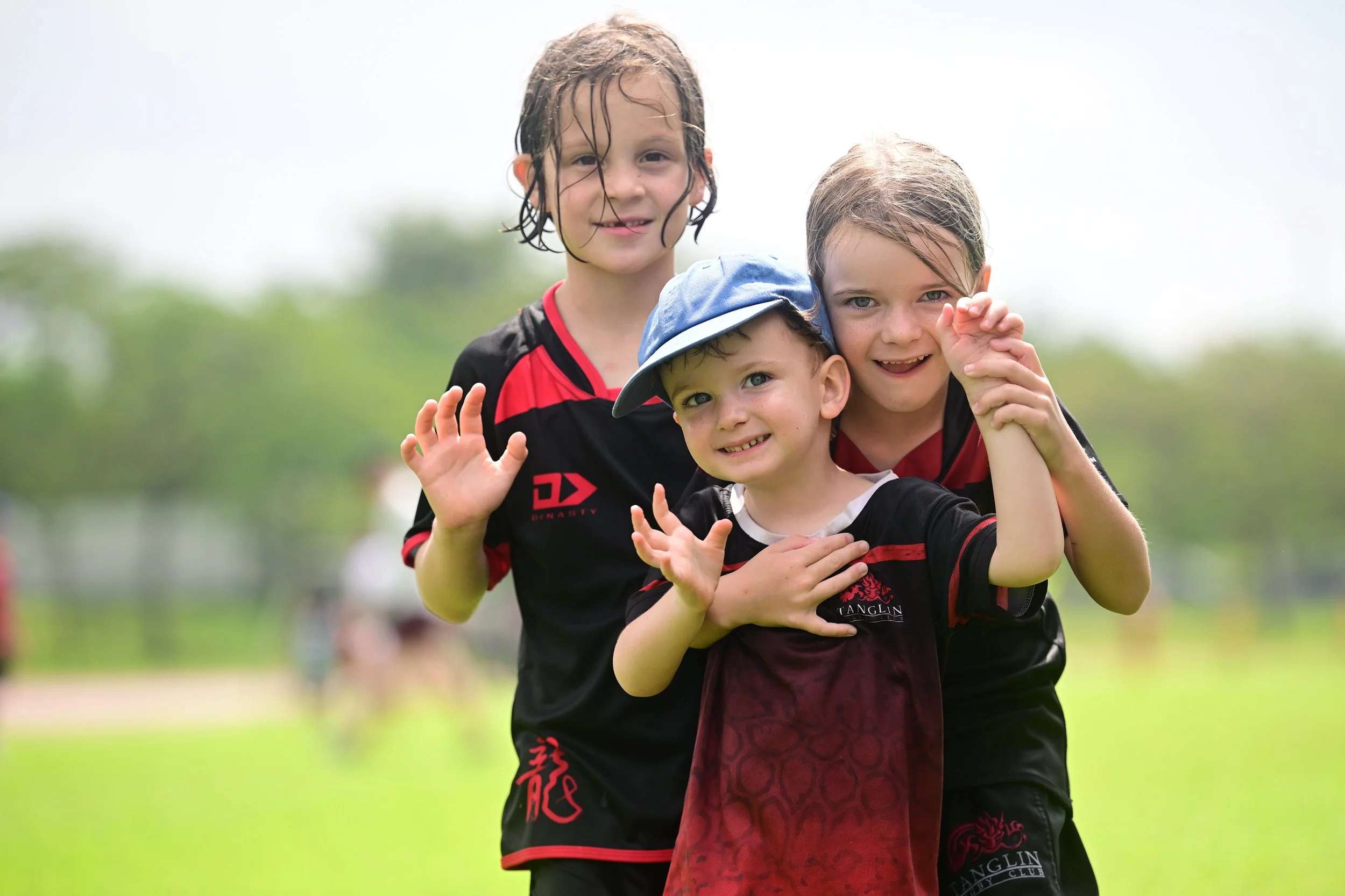 Three children on a grassy field, smiling and posing for the camera, with trees in the background.