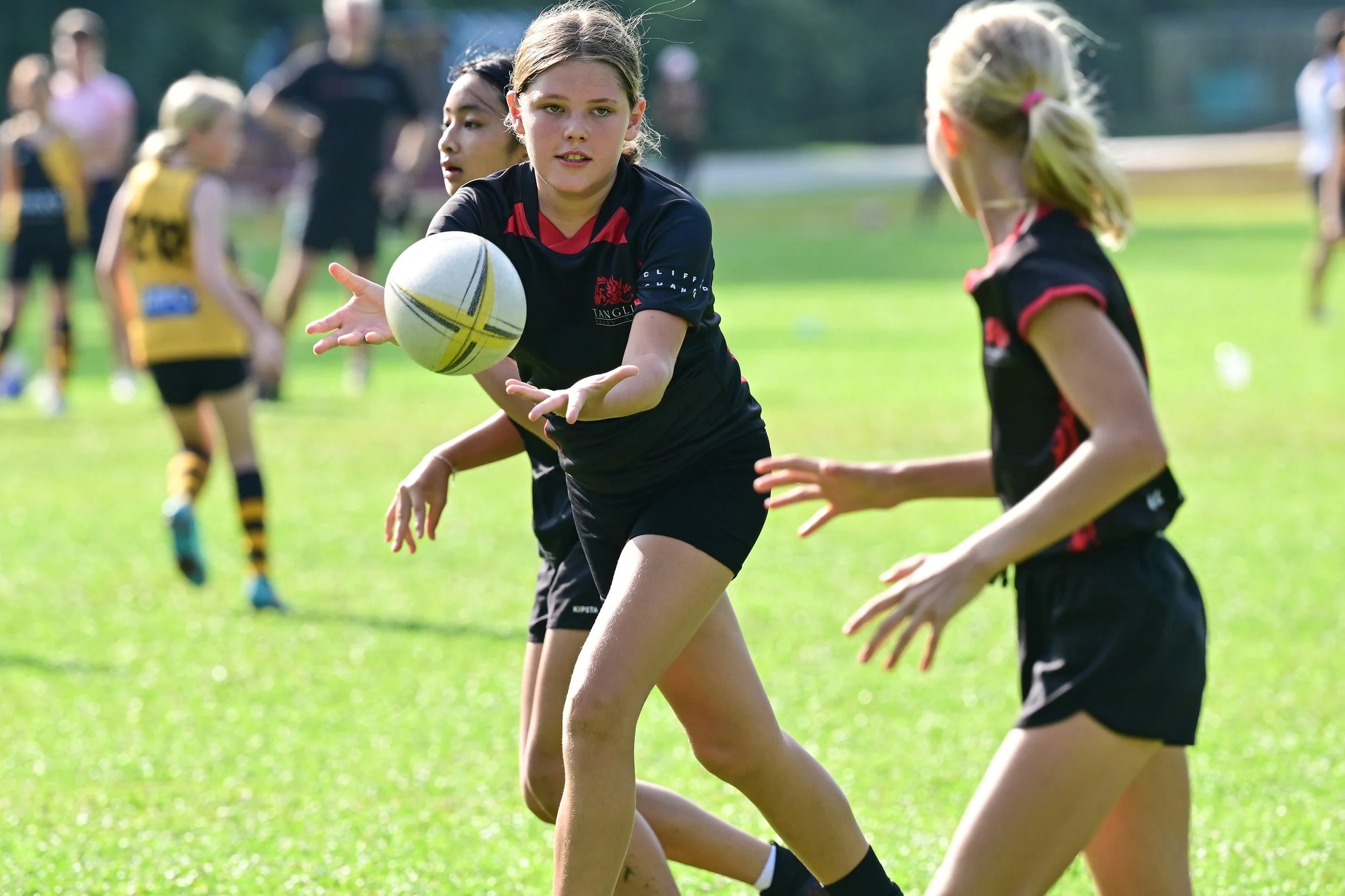 Children playing rugby on a grassy field with one girl passing the ball to a teammate.