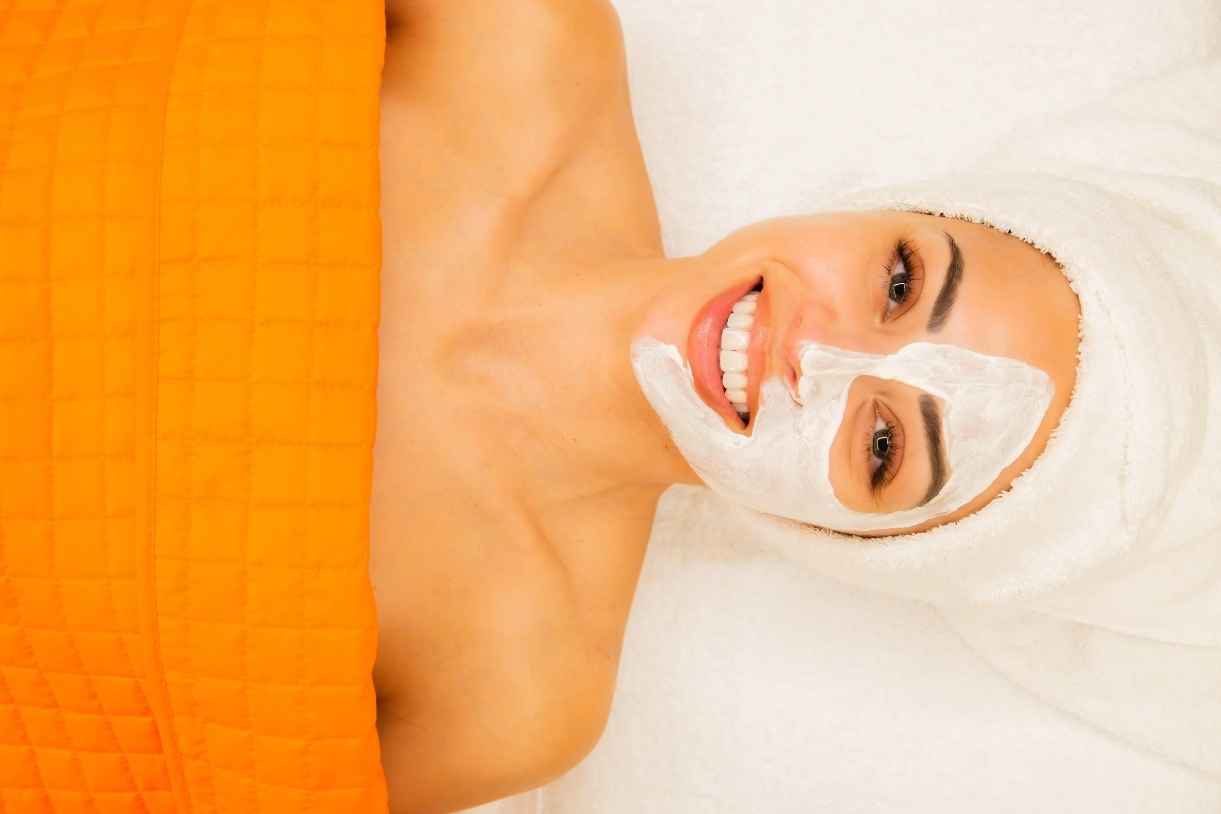 Woman lying on a spa bed with an orange towel, smiling, wearing a towel turban, and a white facial mask.