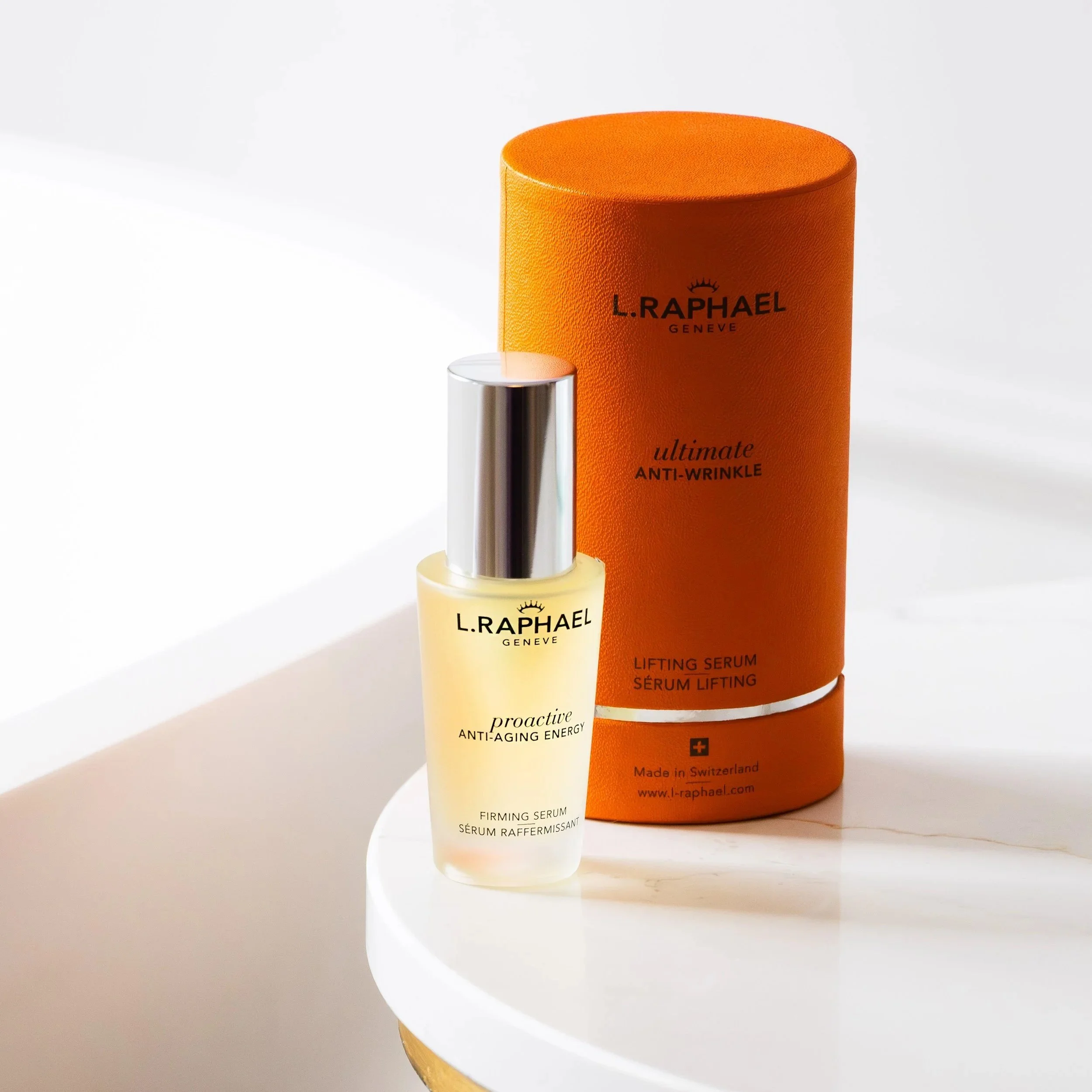 L. Raphael Proactive Anti-Aging Energy firming serum bottle with silver pump next to an orange cylindrical box labeled 'Ultimate Anti-Wrinkle Lifting Serum' on a white circular surface.