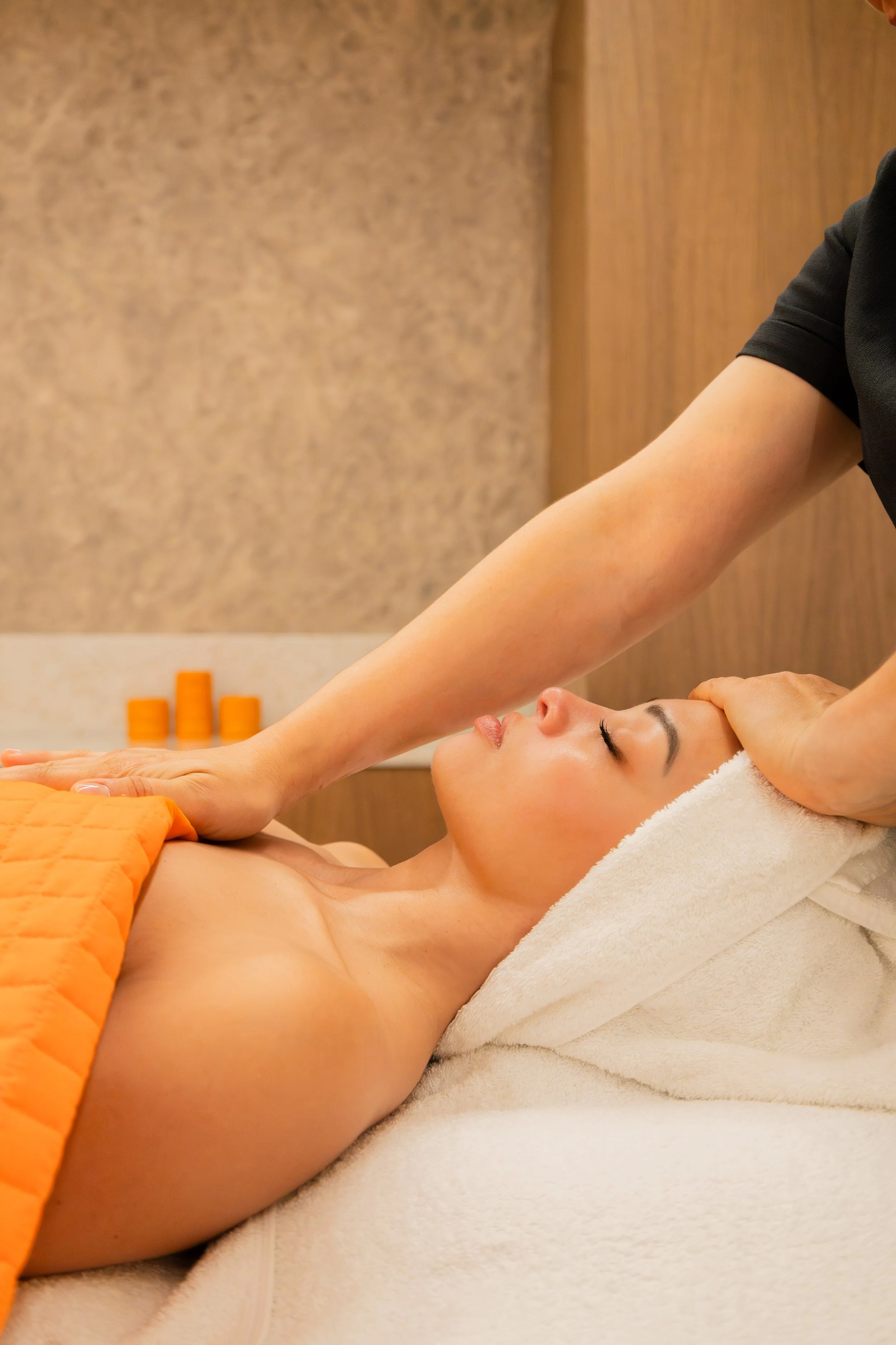 A woman receives a massage at a spa, lying face-up with a towel over her chest.