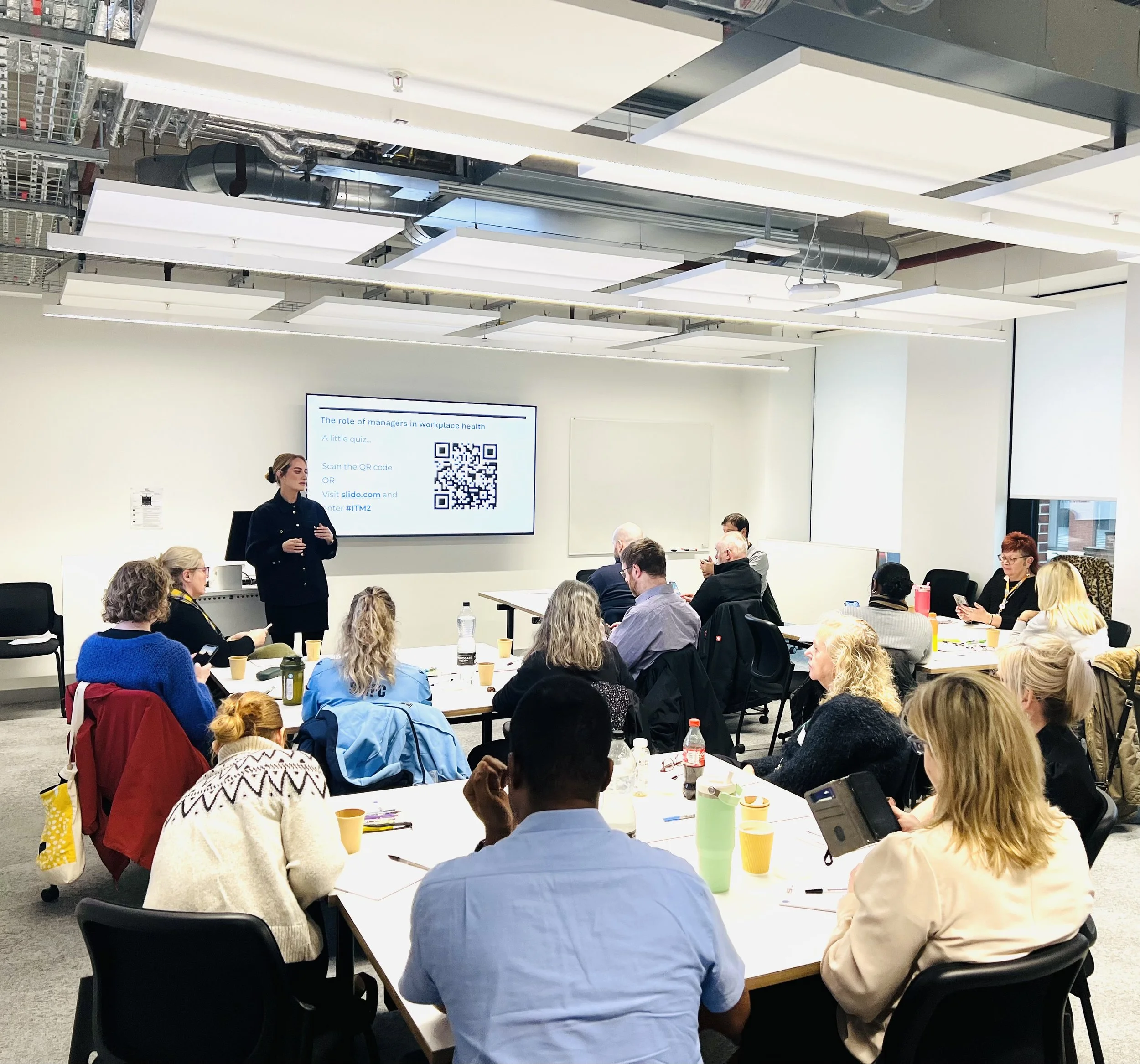 Charlotte on of the Train the Trainers speaking to a group of participants about workplace health and positive psychology, listening sat at tables
