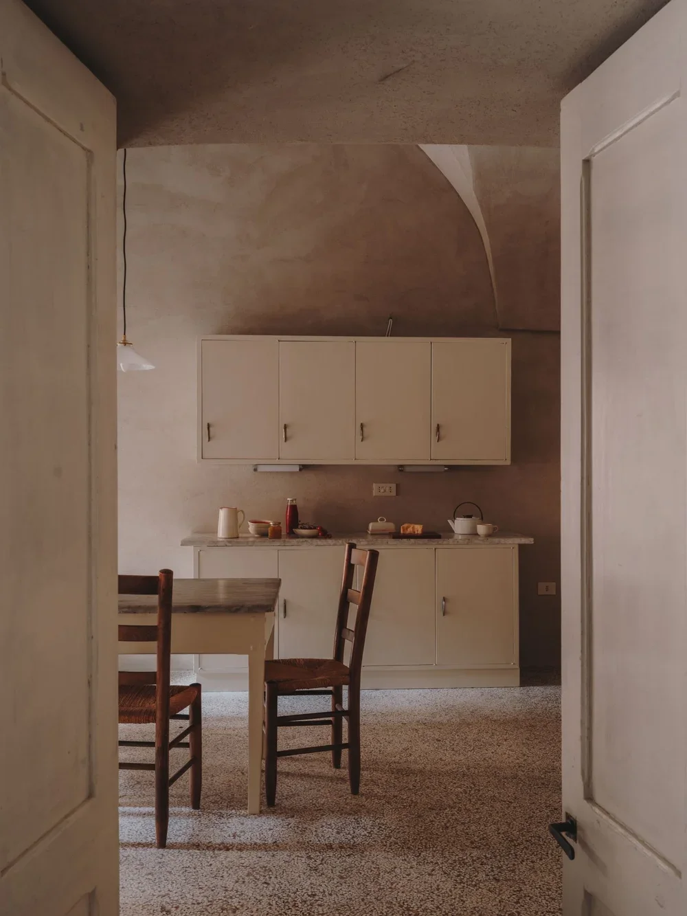 A vintage kitchen with white cabinets, a dining table, and chairs. The room has textured beige walls and ceiling, with a expose light socket hanging from the ceiling. The kitchen counter has a few dishes and kitchenware.