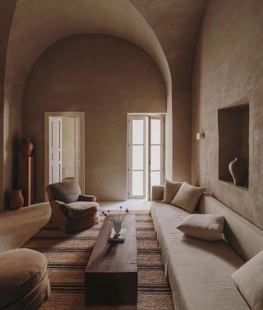 A living room with beige walls and high arched ceiling, featuring a large sofa with throw pillows, a wooden coffee table with a vase of decorative sticks, an armchair, a rug with horizontal stripes, and built-in wall niches with vases. There are double glass doors and a doorway leading to other rooms.