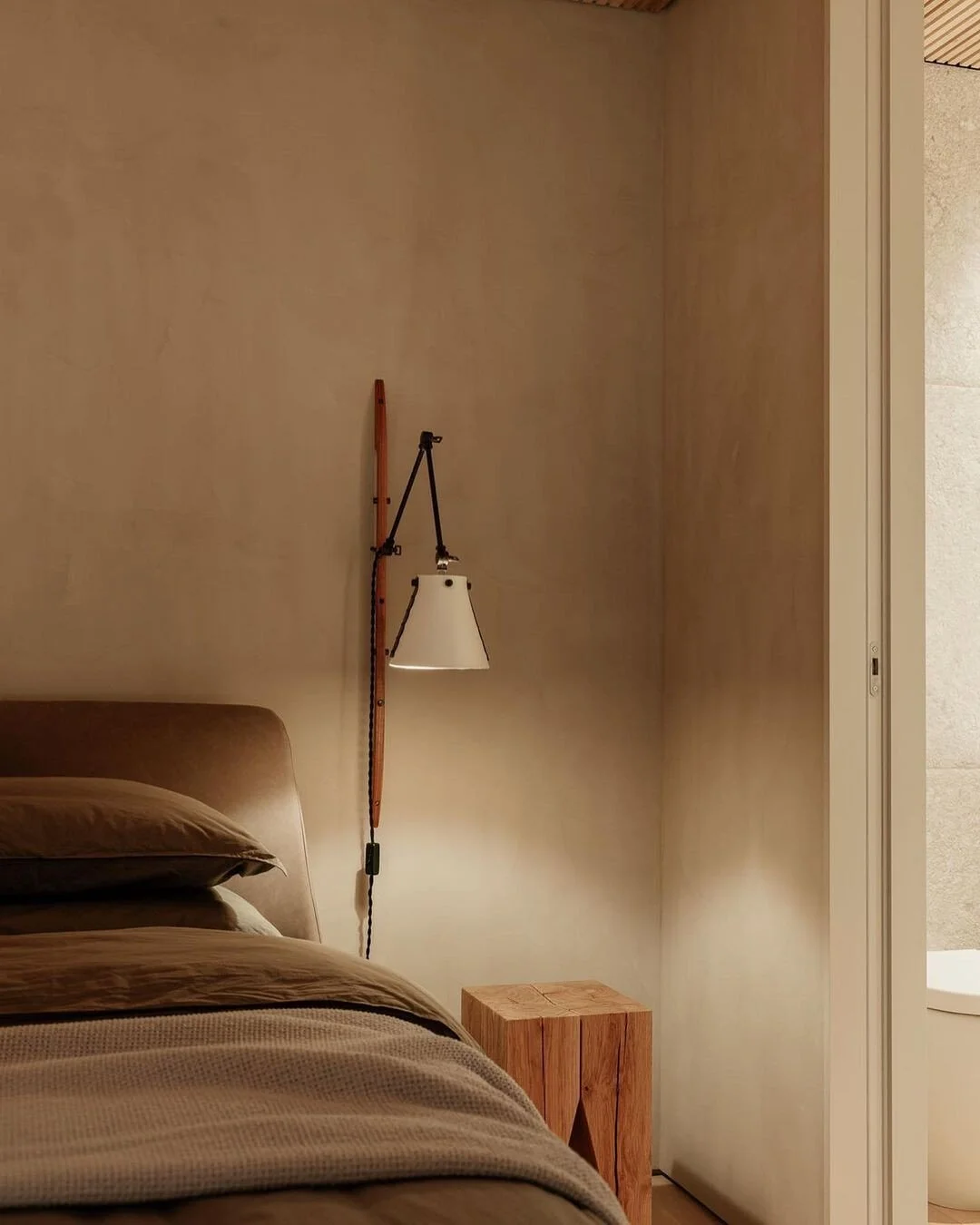 A bedroom with a beige bed, a wooden side table, and a wall-mounted reading lamp with a cone-shaped shade next to a beige wall and an open door leading to a bathroom with beige tiles.