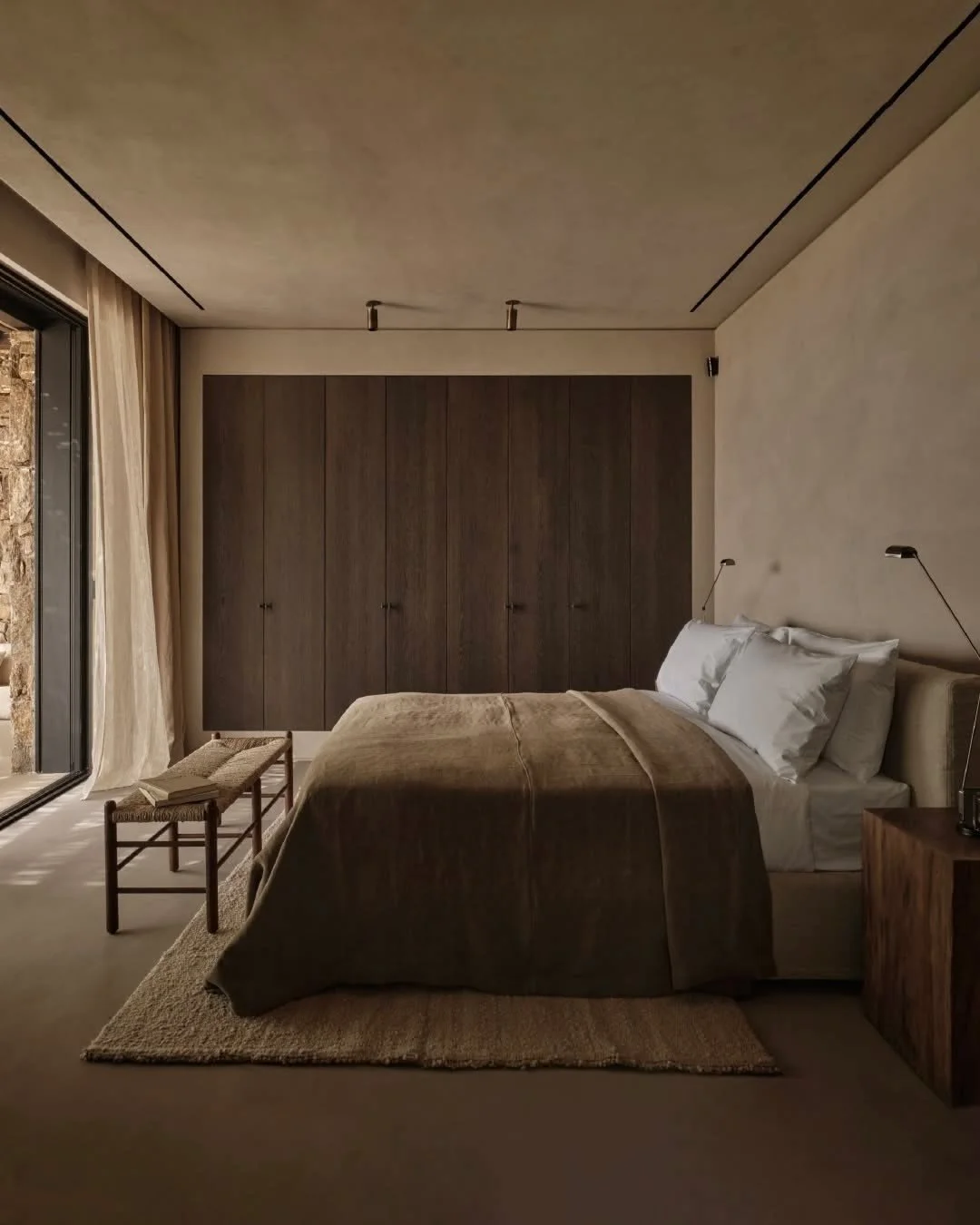 A minimalist bedroom with a bed, bedside tables, reading lamps, a bench at the foot of the bed, and large sliding glass doors with curtains, overlooking a stone wall outside.
