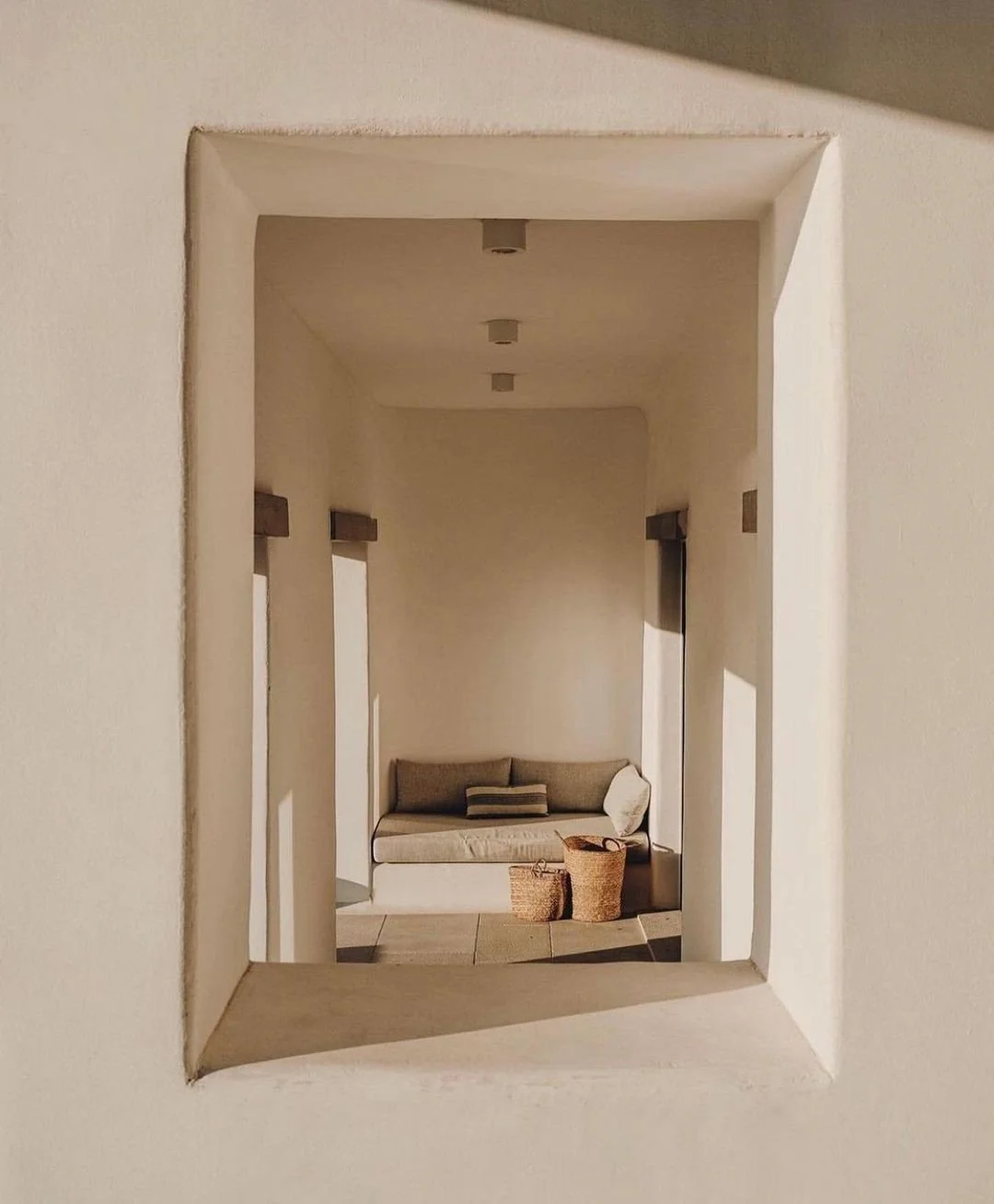 View of a cozy living area seen through a square window, featuring a beige sofa with cushions, two woven baskets, and walls with subtle lighting fixtures.