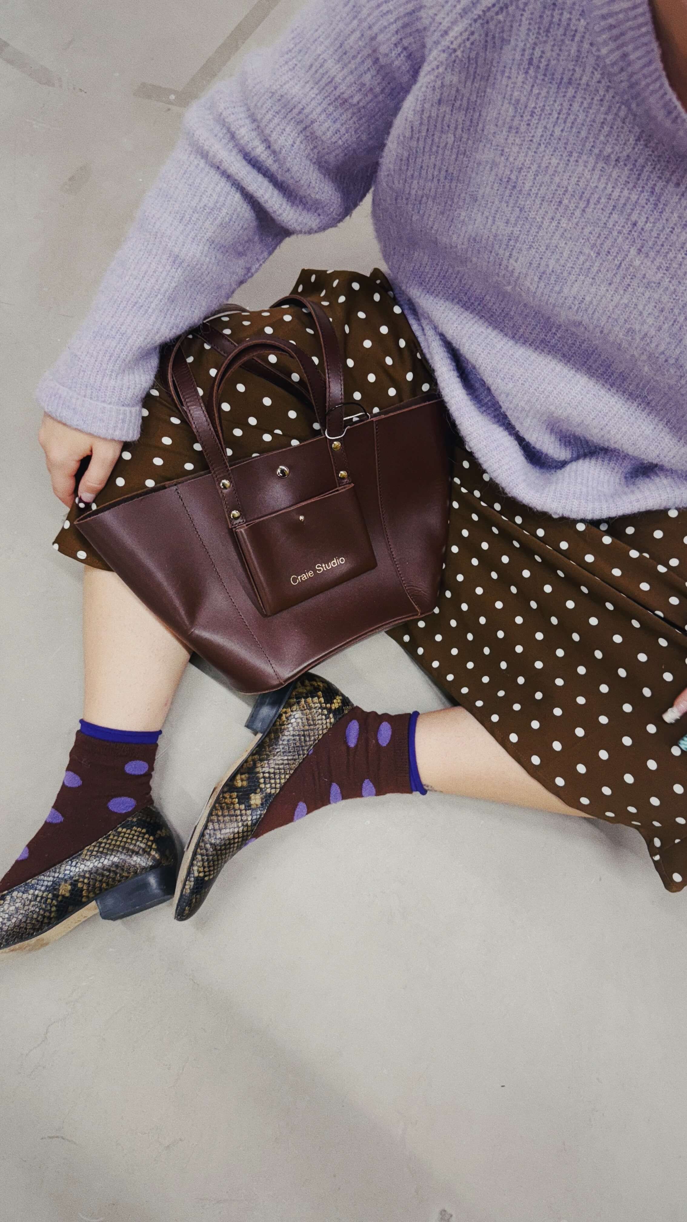 Une personne assise par terre portant une robe marron à pois blancs, un pull violet, des chaussettes violettes à pois marron, des chaussures à talons à motif serpent, un sac en cuir marron et un sac à main marron avec le texte "Craie Studio".