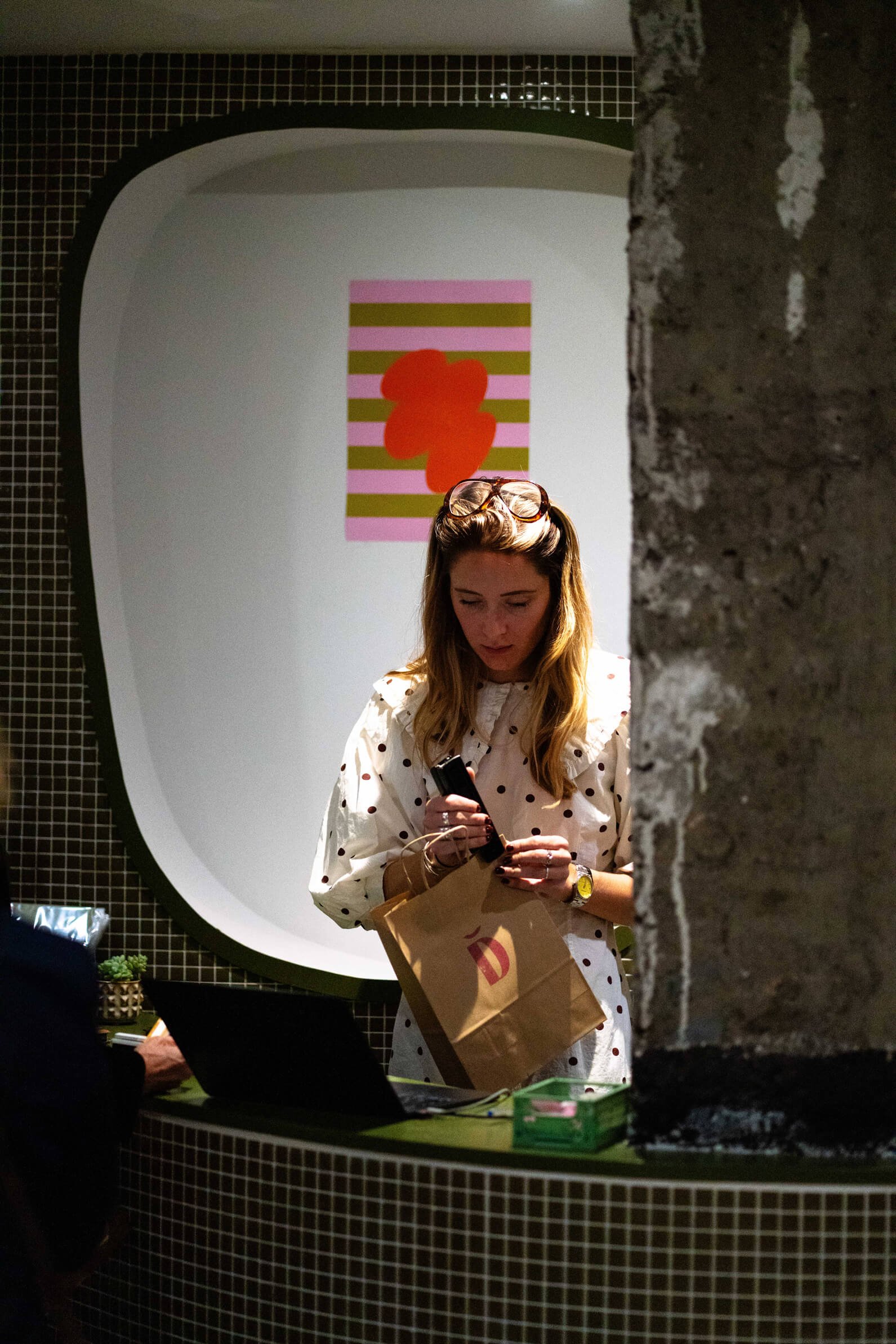 Une femme de "les Dégoteuses" avec des lunettes sur la tête regarde son téléphone, se trouvant derrière un comptoir avec un ordinateur portable et un sac en papier.