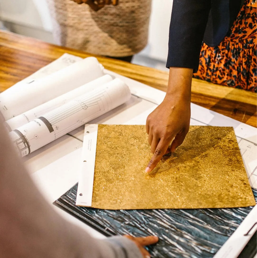 Une personne de "Les Dégoteuses" montrant un échantillon de couleurs dorés ou métalliques lors d'une réunion ou présentation. Sur la table, il y a des plans, des papiers et des rouleaux de schémas.