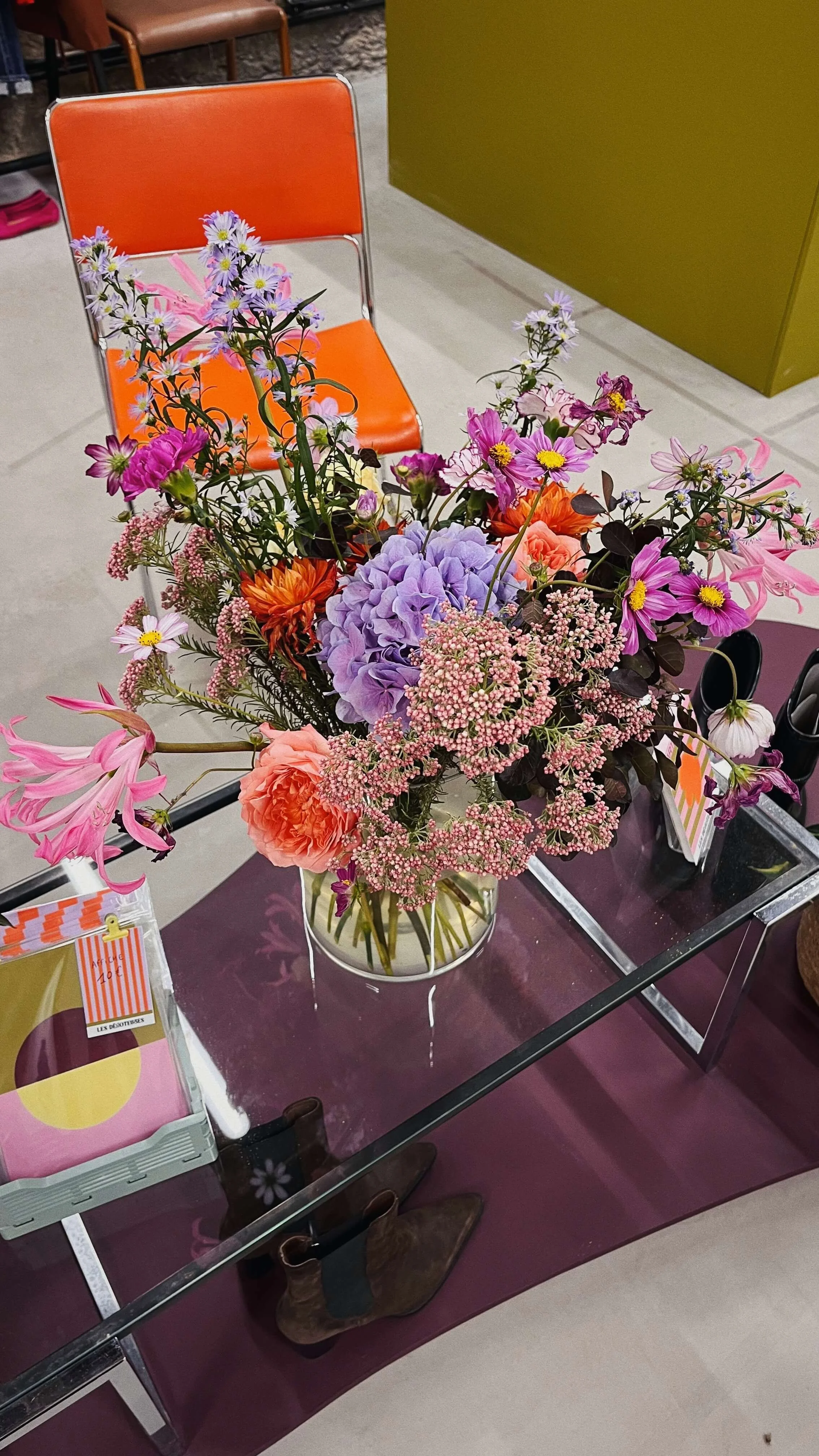 Un bouquet de fleurs colorées comprenant des hortensias violets, des marguerites roses, des roses orange et des fleurs de différentes tailles dans un vase en verre sur une table en verre dans la boutique "les Dégoteuses".
