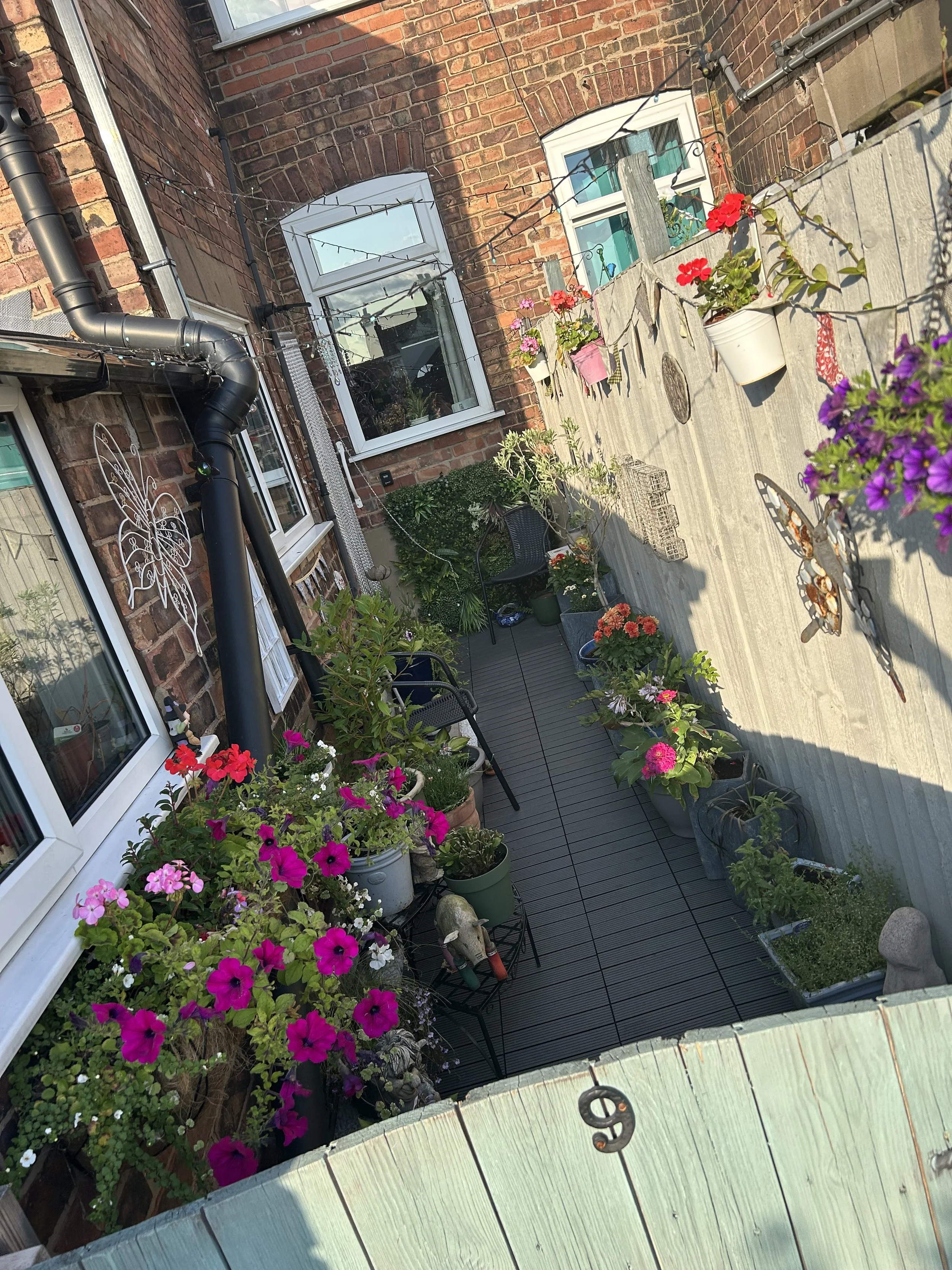 A small backyard patio with potted flowering plants, benches, a wooden fence decorated with butterflies and hanging baskets, and brick house walls with windows and mesh fairy lights.