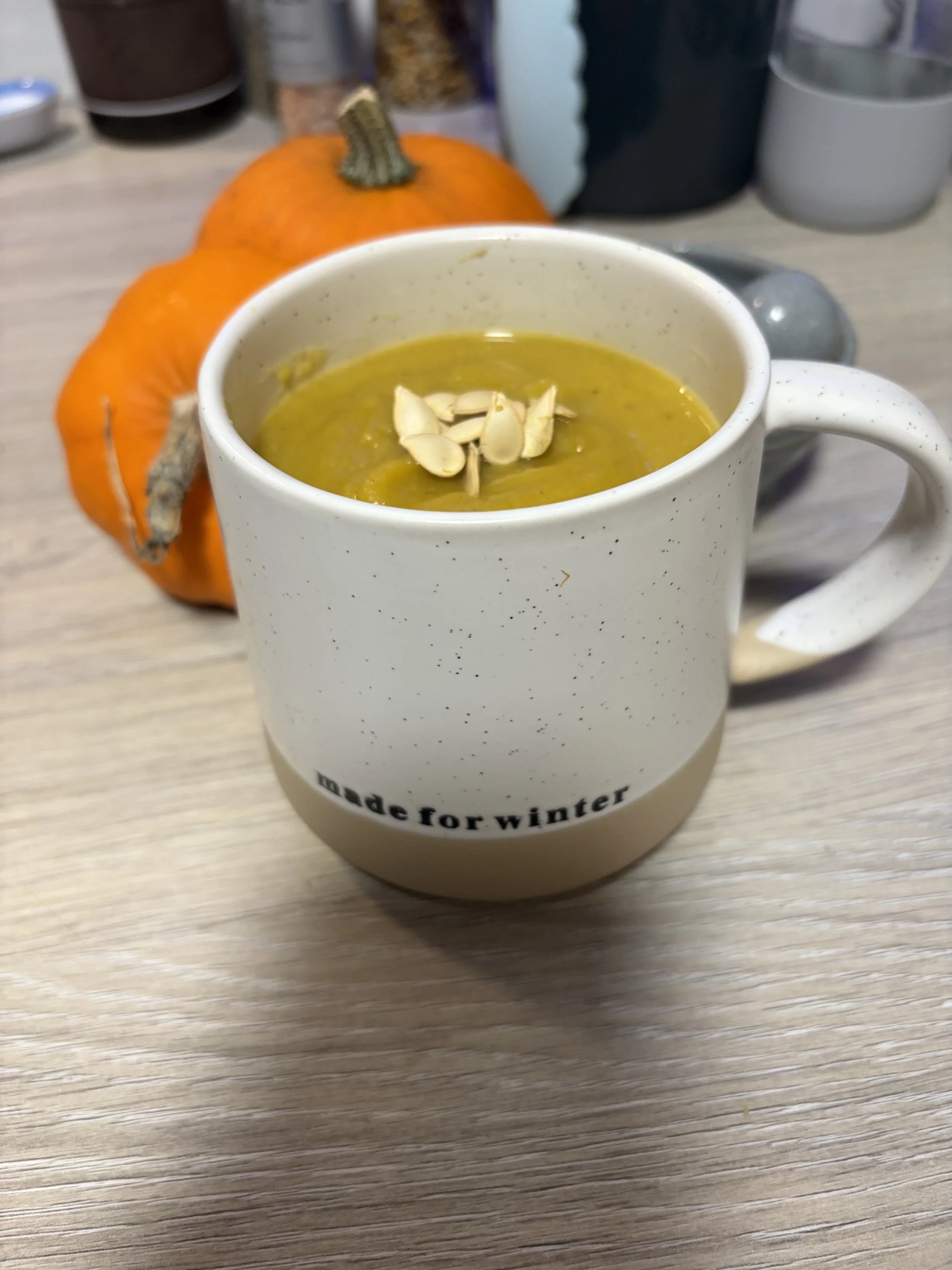 A mug of pumpkin soup topped with pumpkin seeds, placed on a wooden table with two small pumpkins and blurred containers in the background.