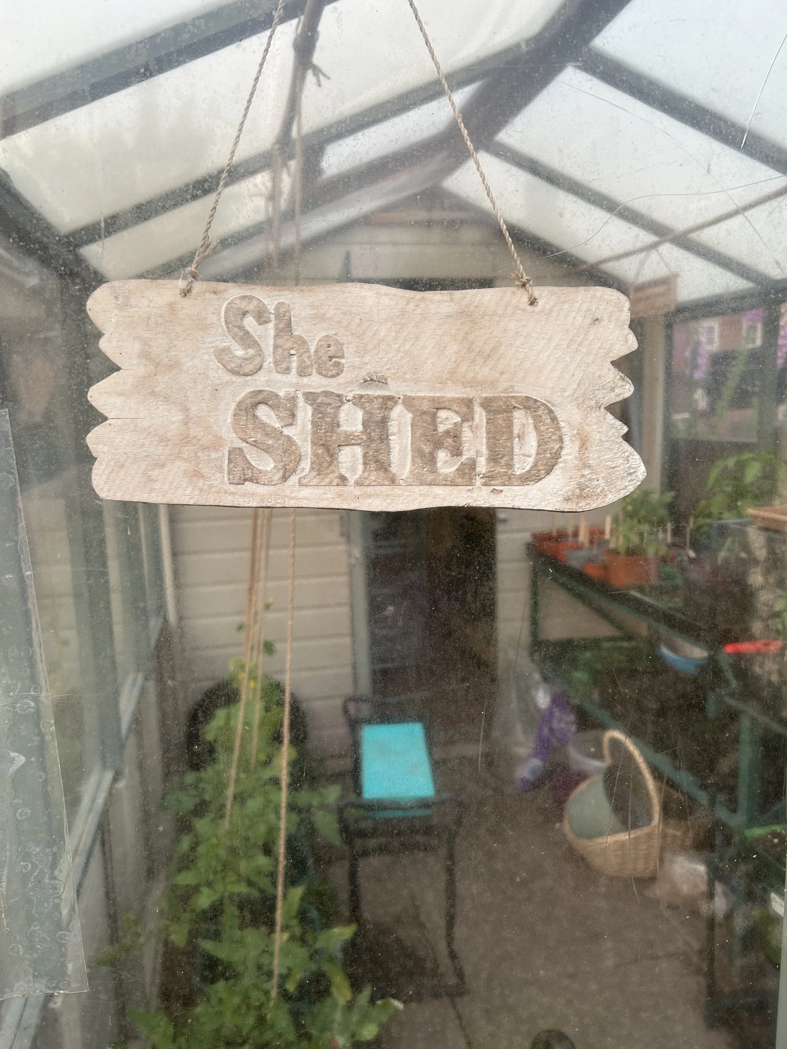 Wooden sign with engraved text reading 'She SHED' hanging inside a greenhouse.