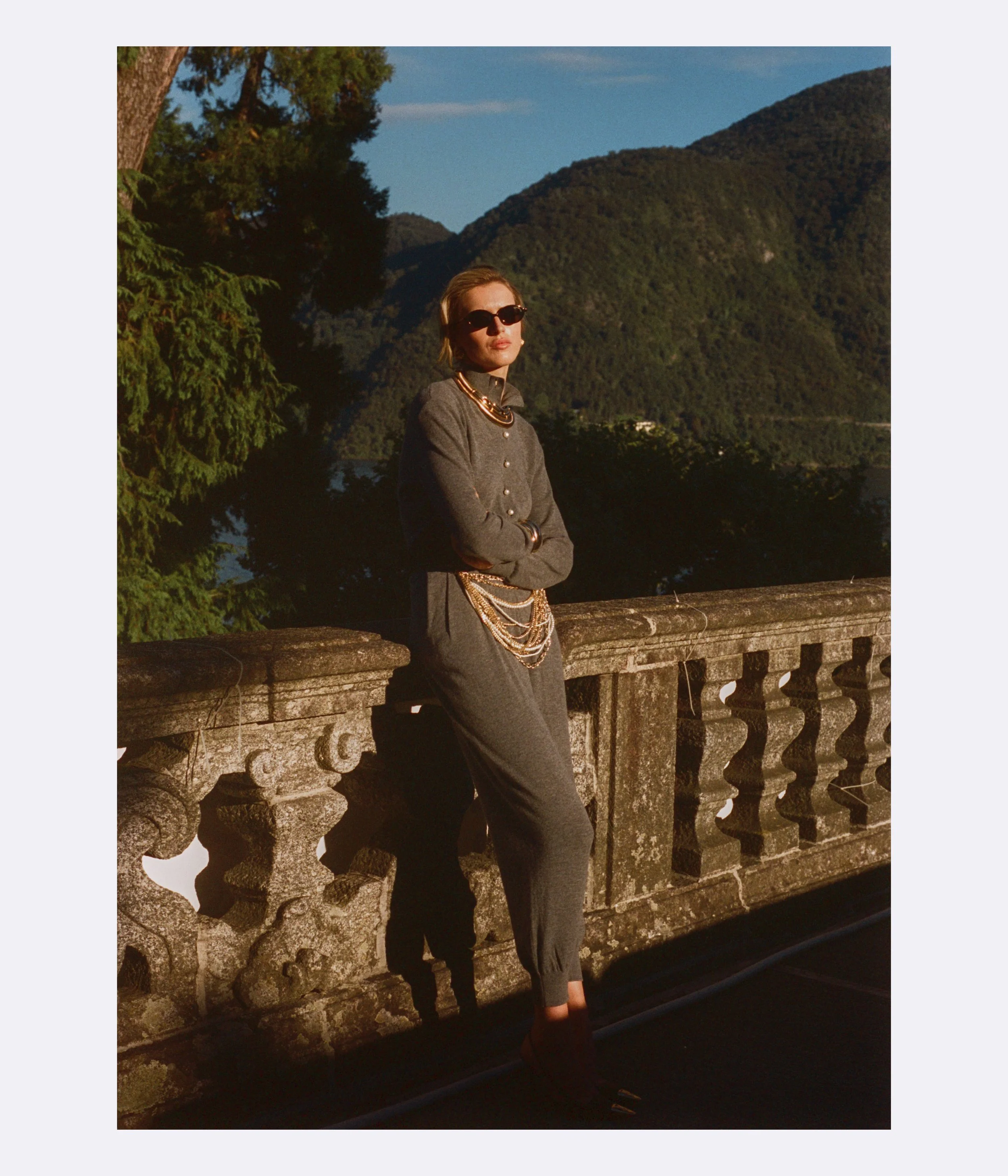 A woman in sunglasses and dark gray outfit with layered jewelry, leaning against a stone railing outdoors during sunset, with green trees and mountain landscape in the background.