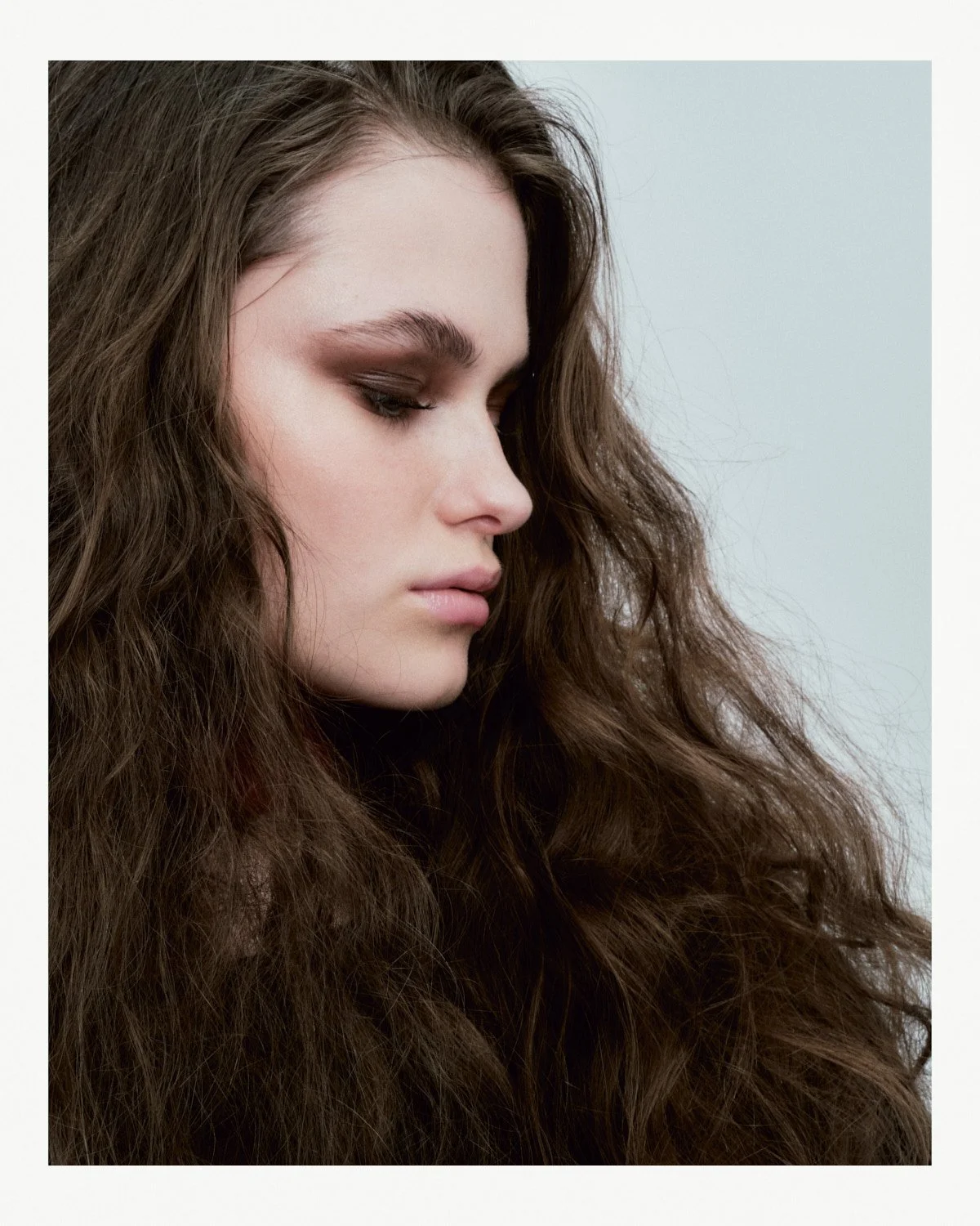 Close-up of a young woman with long, wavy brown hair and makeup, looking to the side with a neutral expression.