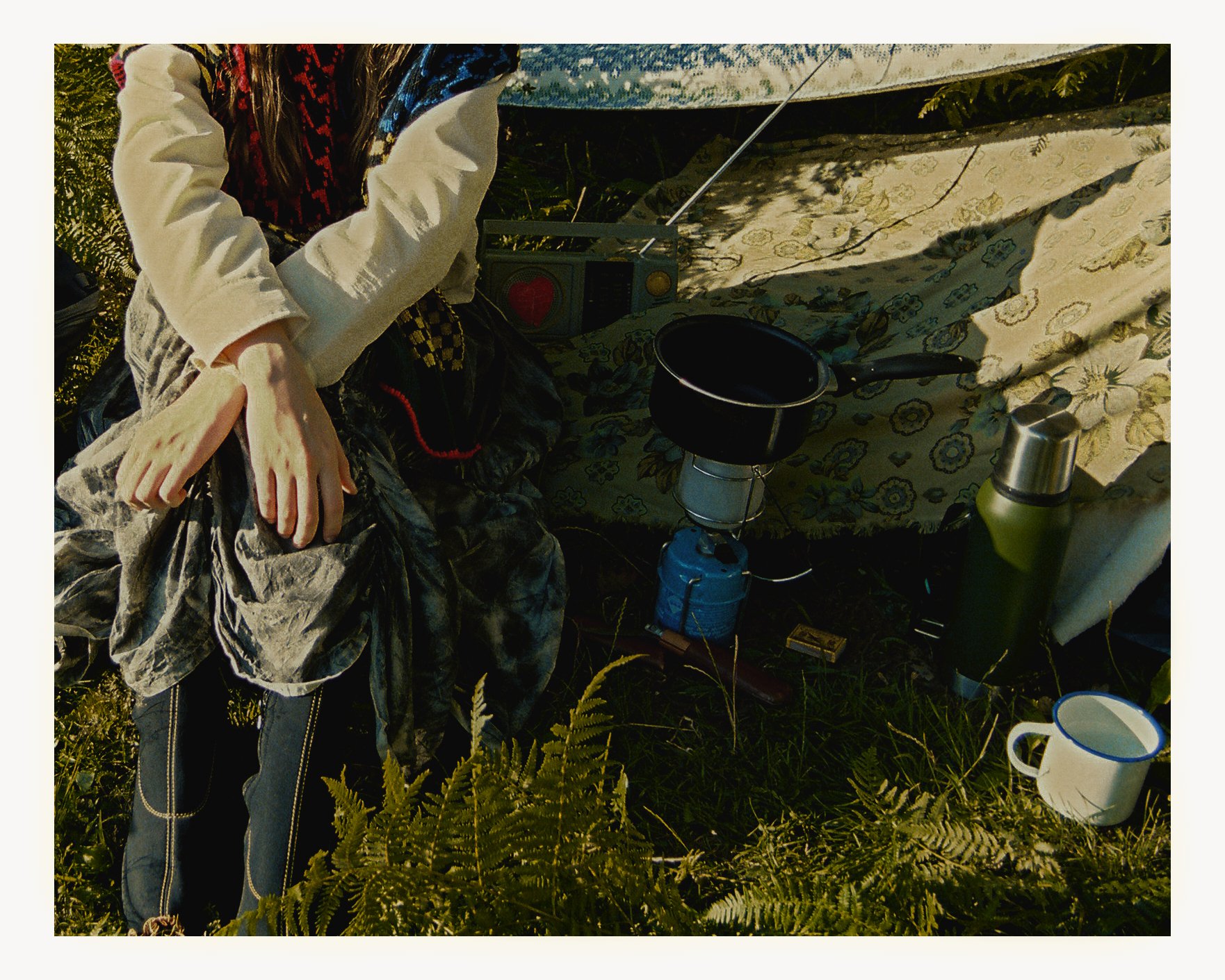 A figure of a mannequin or doll sitting outdoors among grass and ferns, surrounded by camping gear including a camping stove, a pot, a water bottle, a mug, and a speaker, with a floral-patterned blanket or cloth behind it.