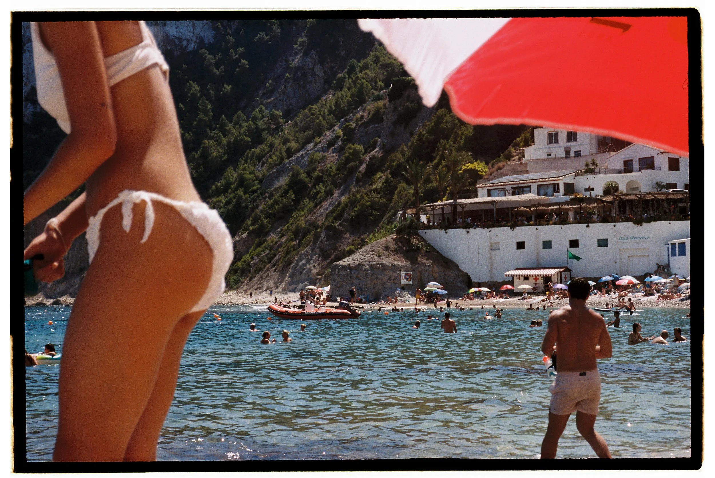 Beach scene with people swimming and sunbathing, hillside buildings, and umbrellas, viewed from under a red and white umbrella.