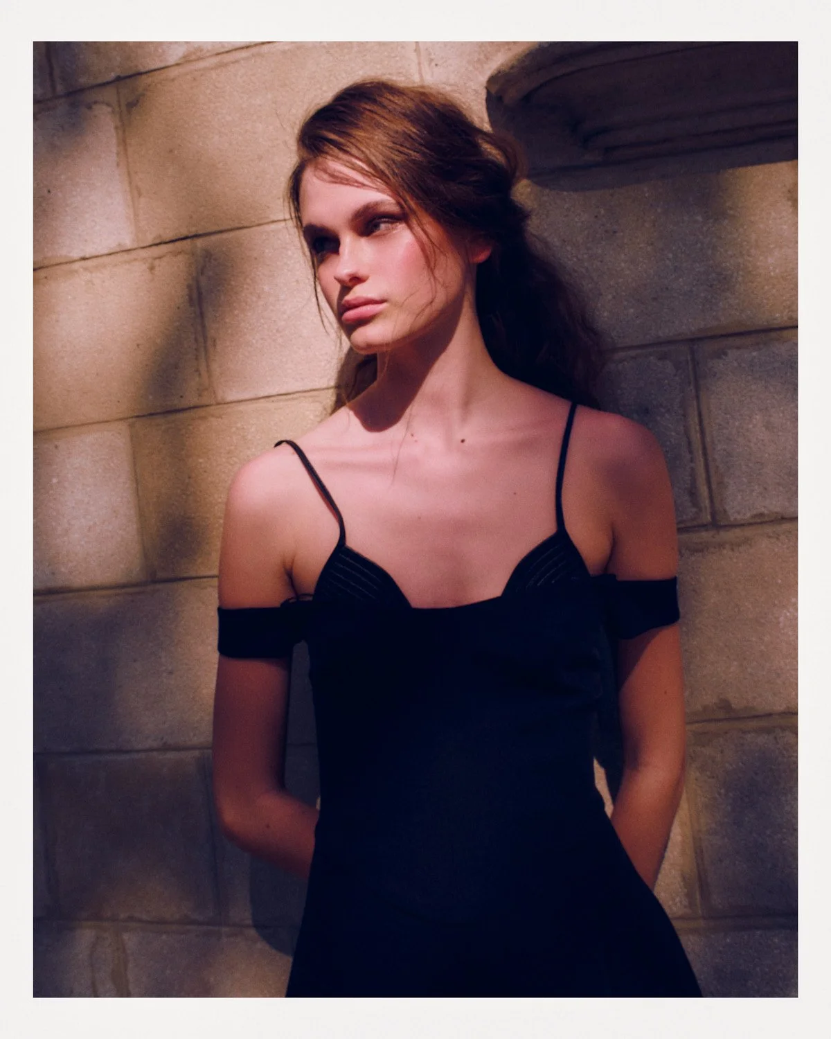 A young woman with brown hair and fair skin, wearing a black dress with thin straps, standing against a stone wall with a column to her right, looking to her left.