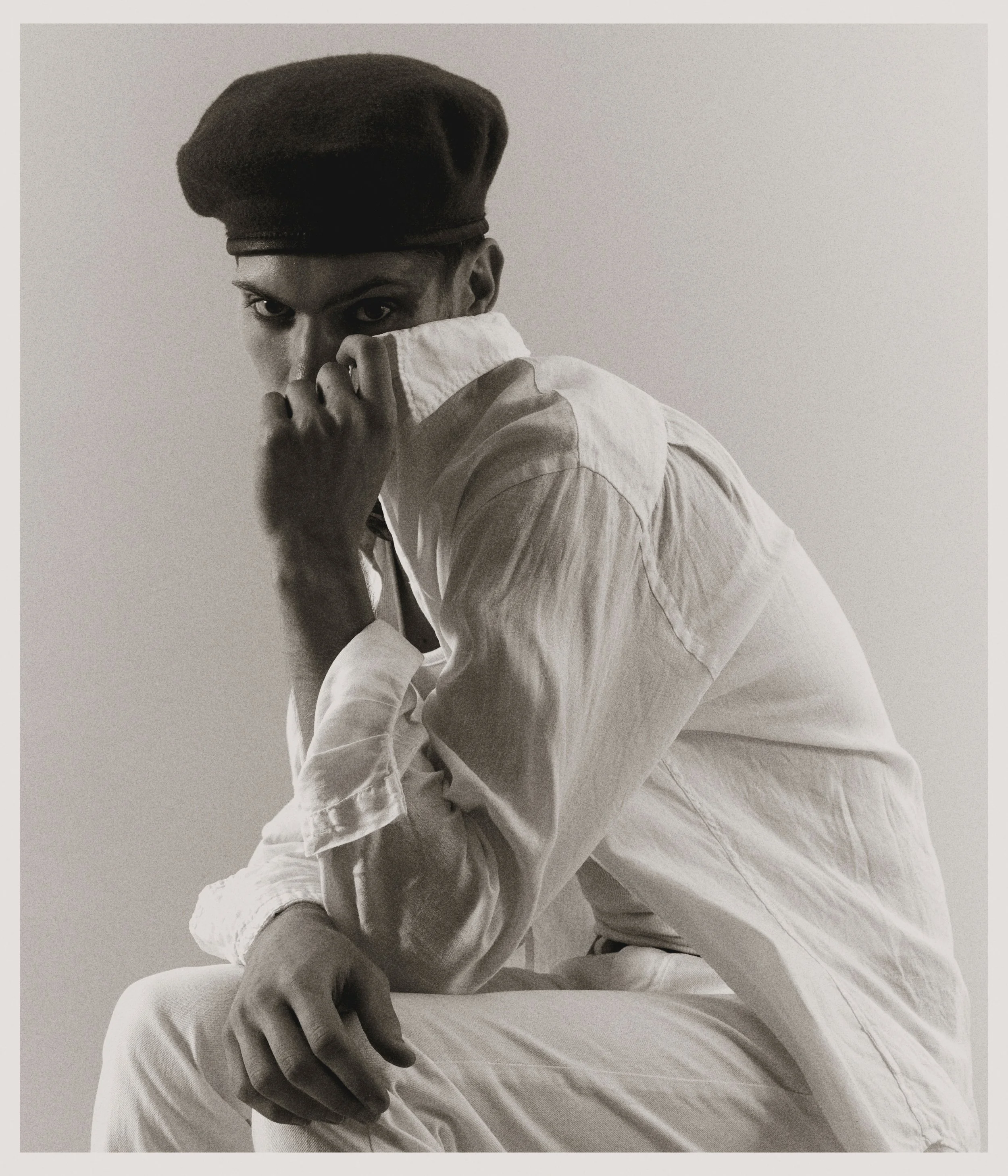 Black and white photo of a person wearing a beret and a light-colored shirt, sitting and partially covering their face with their hand, looking intently at the camera.